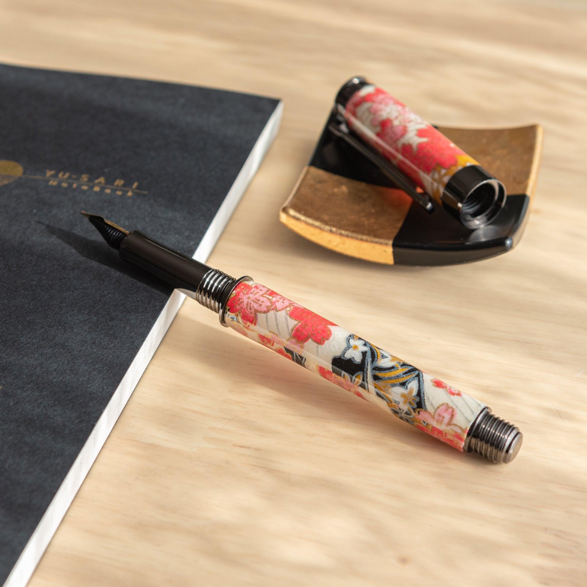 Open fountain pen with floral cherry blossom design resting on a black notebook, with its cap placed on a gold-accented pen holder nearby.