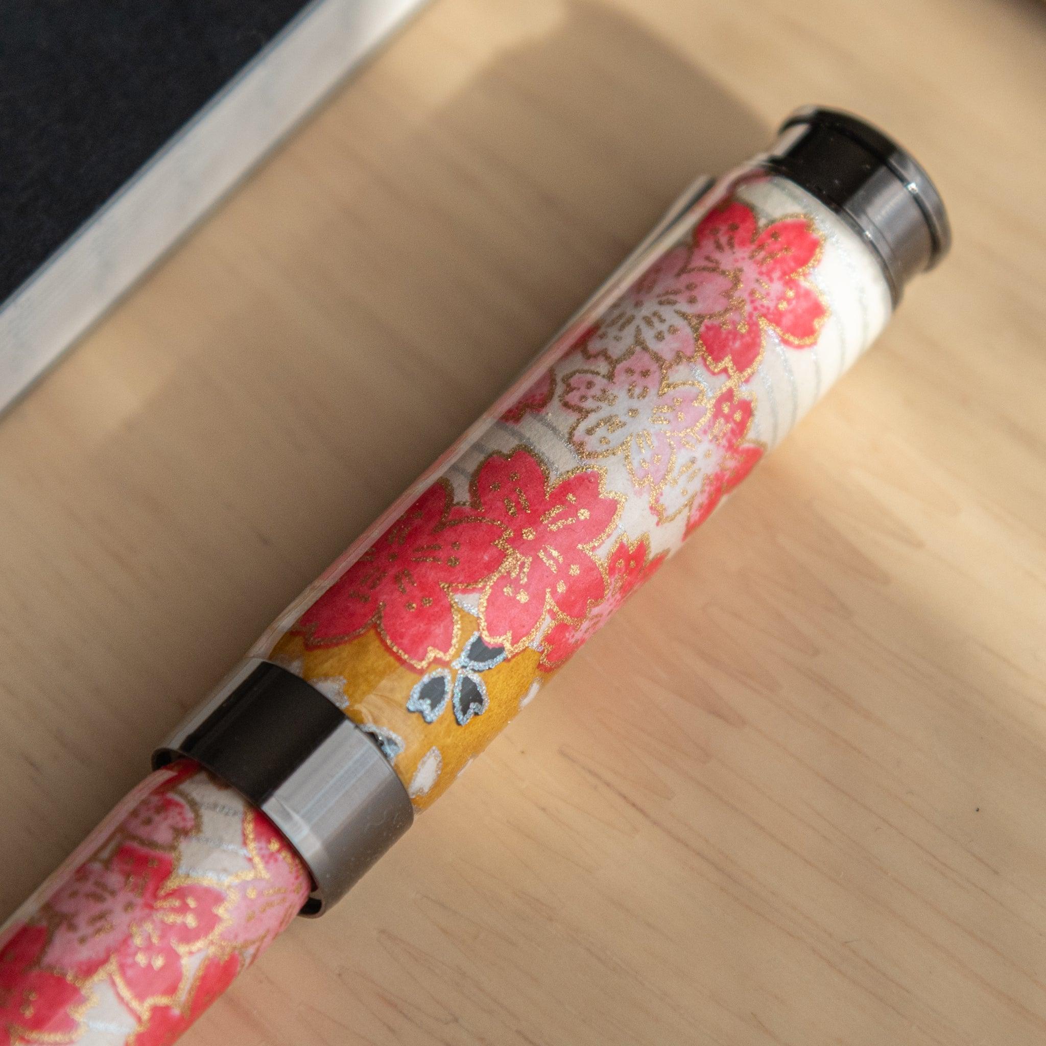 Close-up of a fountain pen cap, decorated with vibrant cherry blossom patterns and gold accents on a washi-style background.