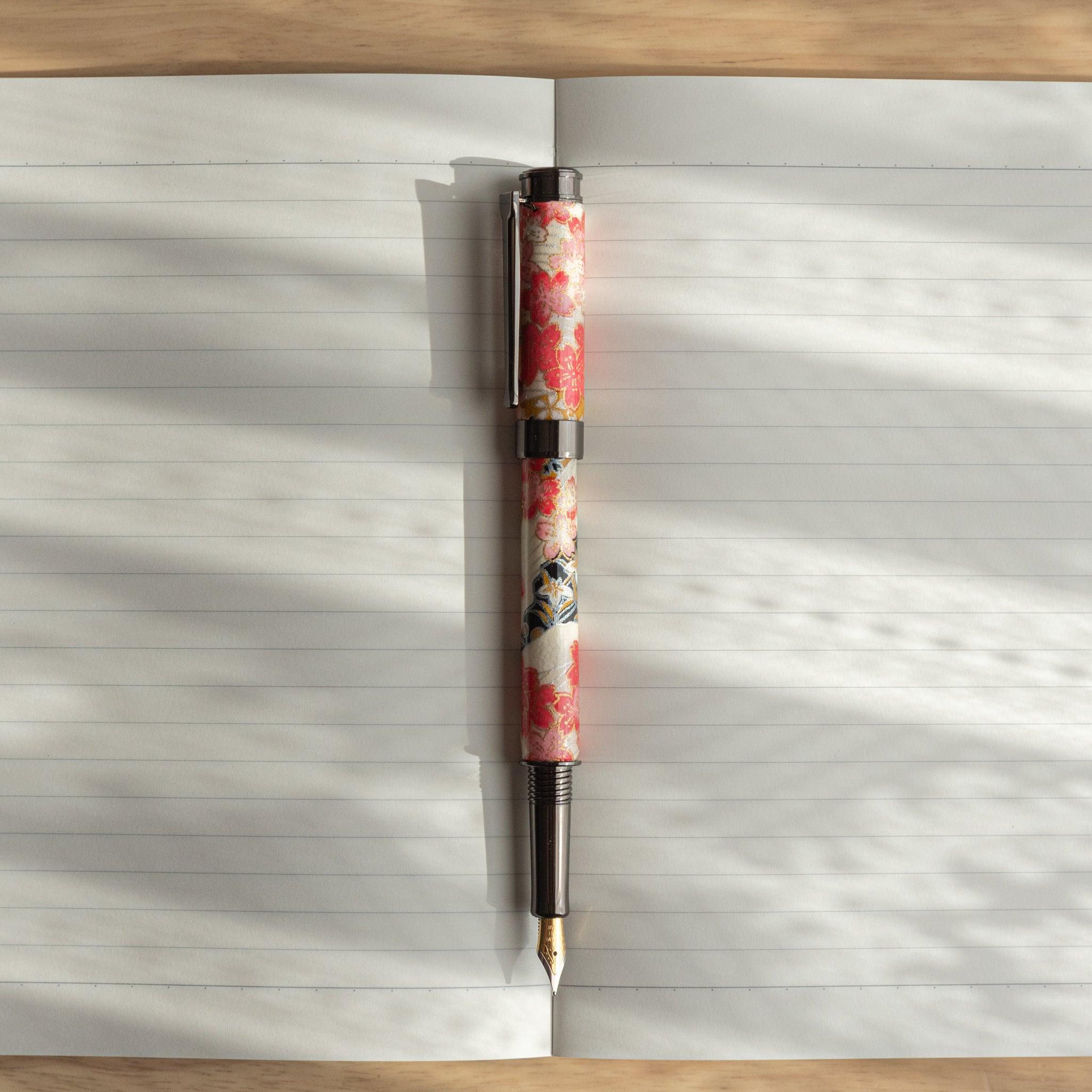 Floral fountain pen with cherry blossom motifs resting on an open notebook with lined pages, illuminated by soft natural light.