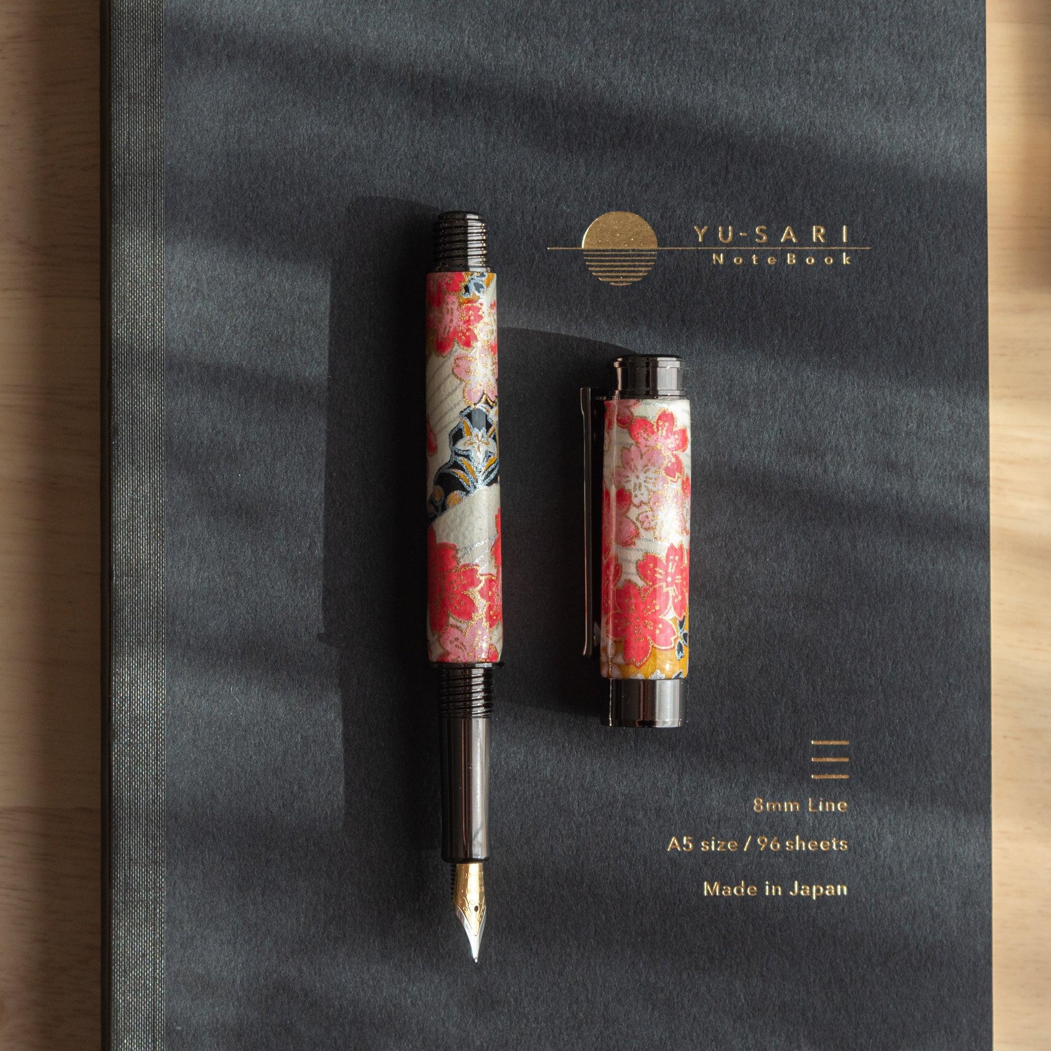 Floral-patterned fountain pen and its cap placed on a black "Yu-Sari" notebook with gold text, illuminated by natural light.