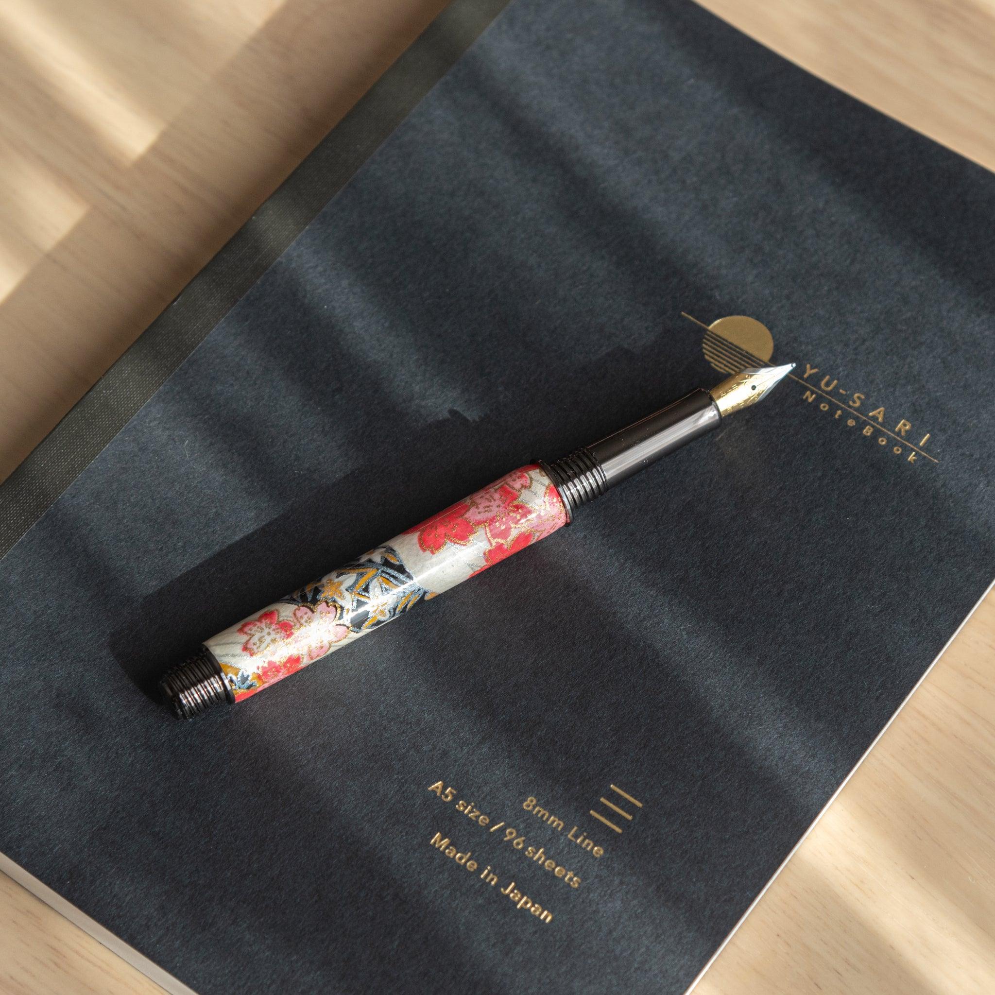 Fountain pen with cherry blossom design and gold nib resting diagonally on a black "Yu-Sari" notebook, featuring gold-embossed details.