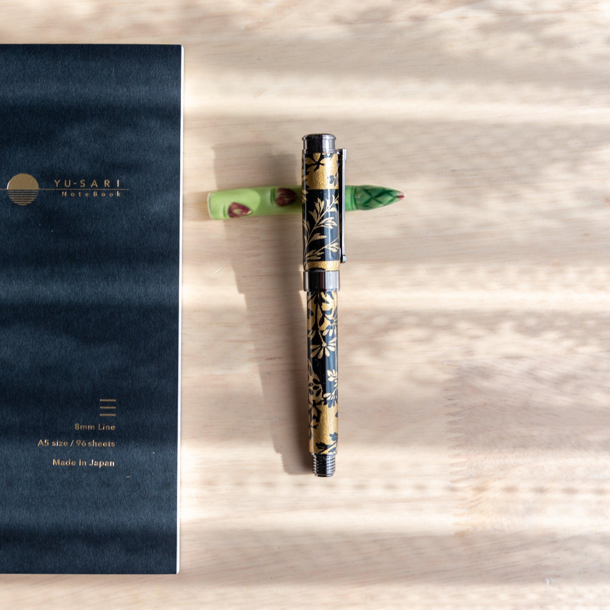 A handmade green ceramic pen rest shaped as an asparagus, holding a black and gold fountain pen. Part of traditional Japanese stationery items.