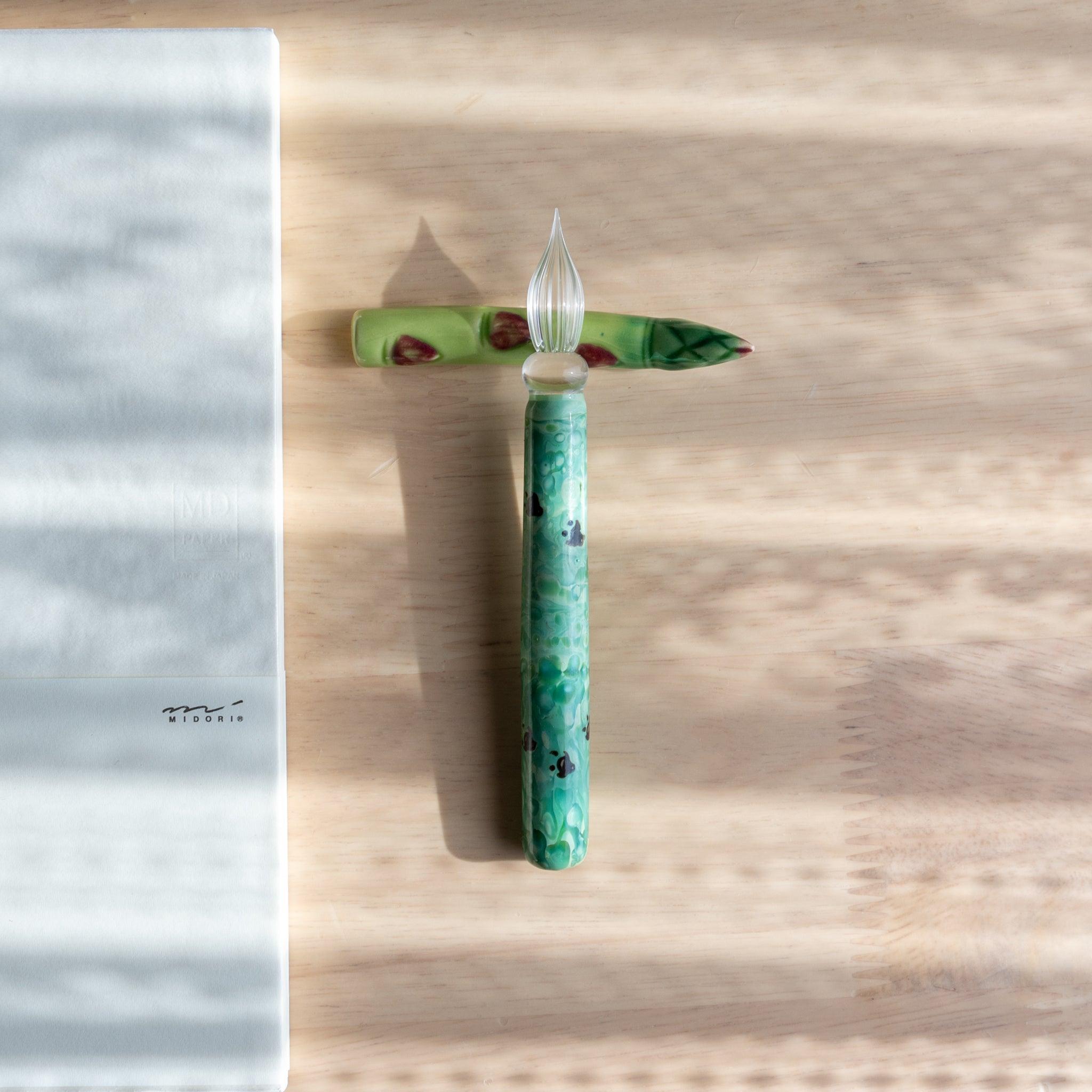 A handmade green ceramic pen rest shaped as an asparagus, holding a green glass dip pen. Part of traditional Japanese stationery items.