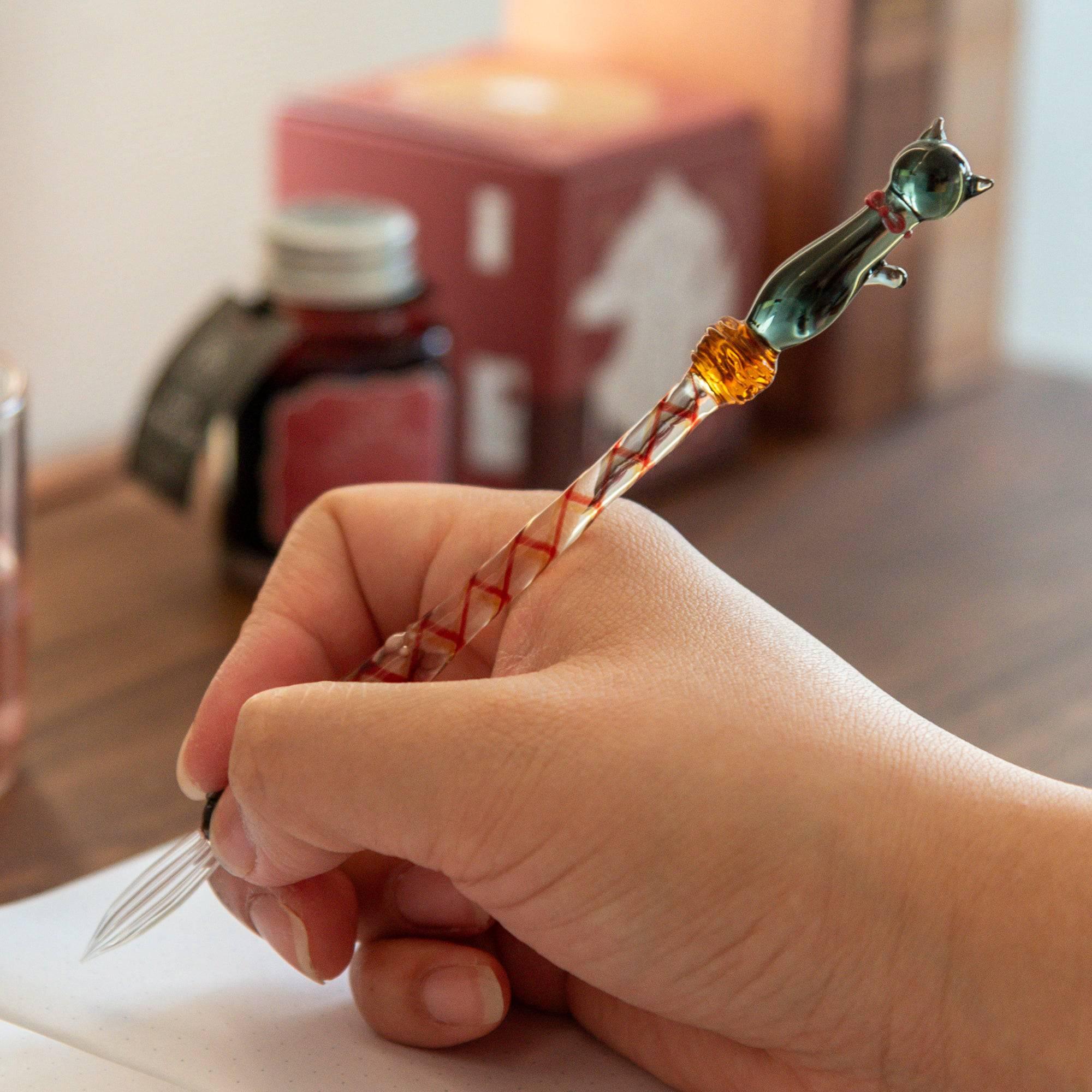 Hand holding a glass pen with a black cat design, writing in a notebook, showcasing the pen's smooth and elegant use.