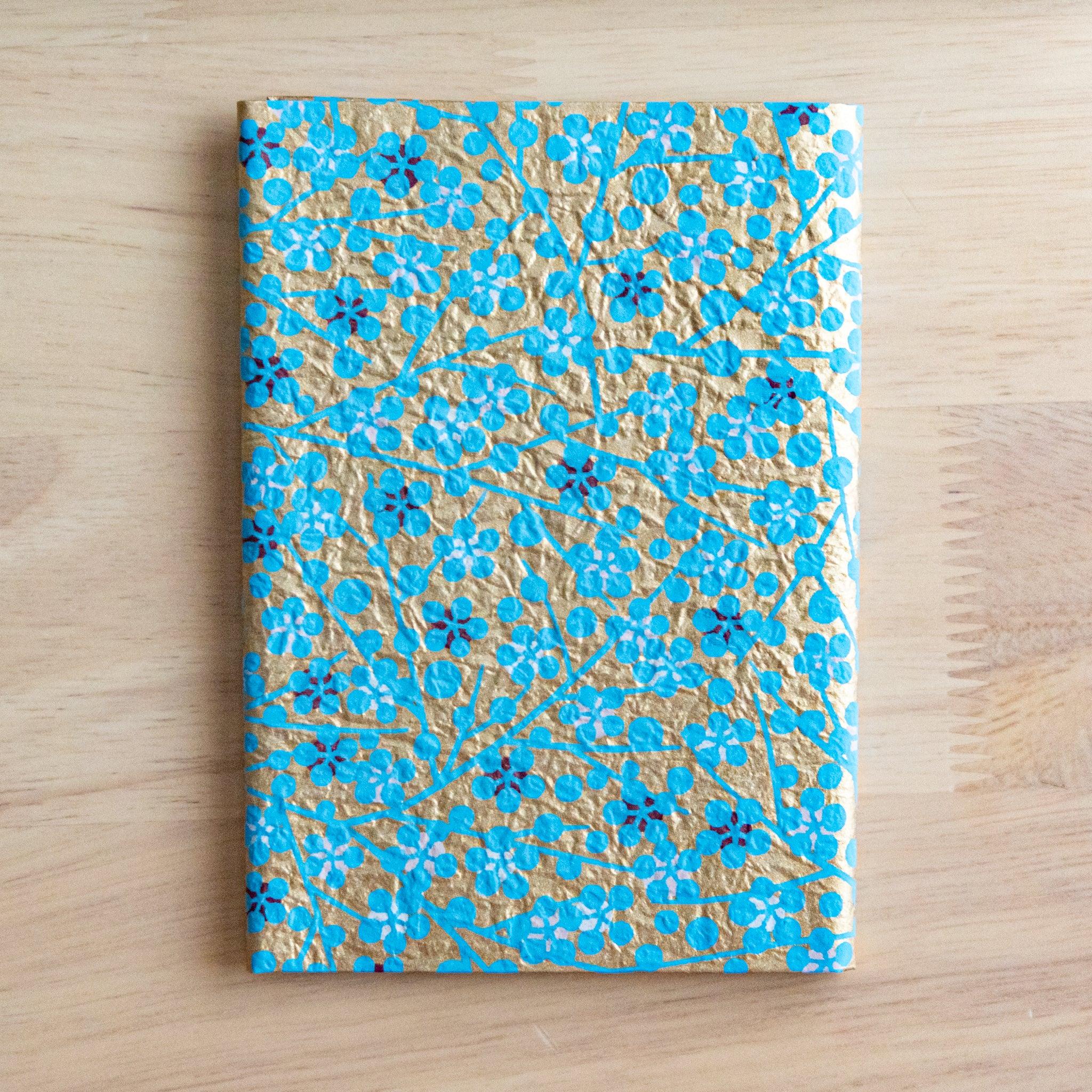 A small notebook with a gold and blue floral pattern on its cover, placed on a light wooden surface.