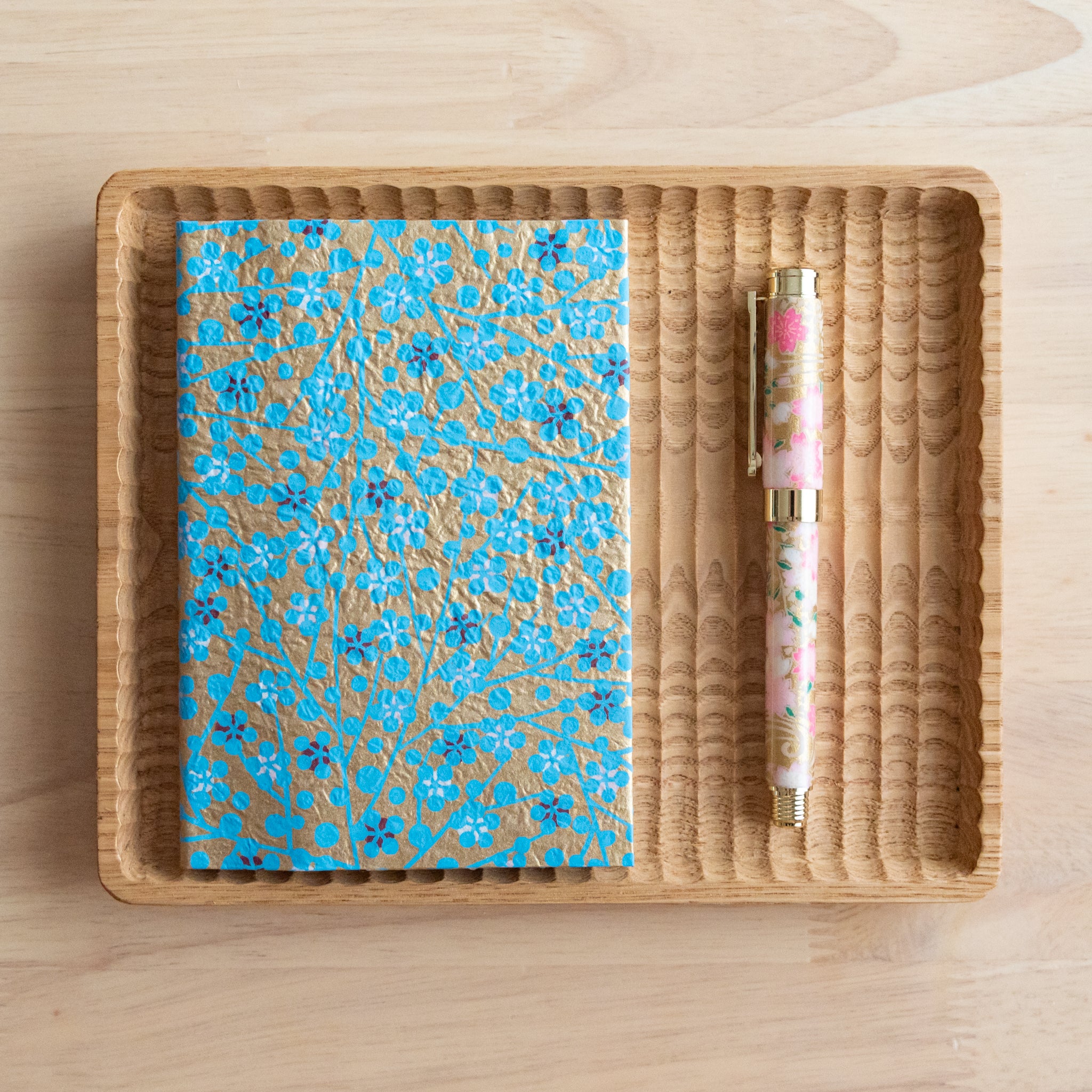 A gold and blue floral patterned notebook and a pink floral pen arranged on a wooden tray on a light wooden surface.