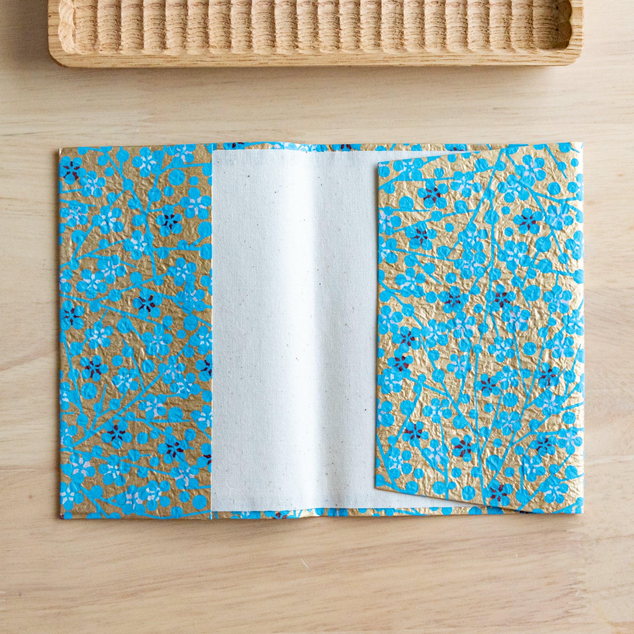 An open notebook cover with a gold and blue floral pattern, revealing its inner white lining, placed on a wooden surface.