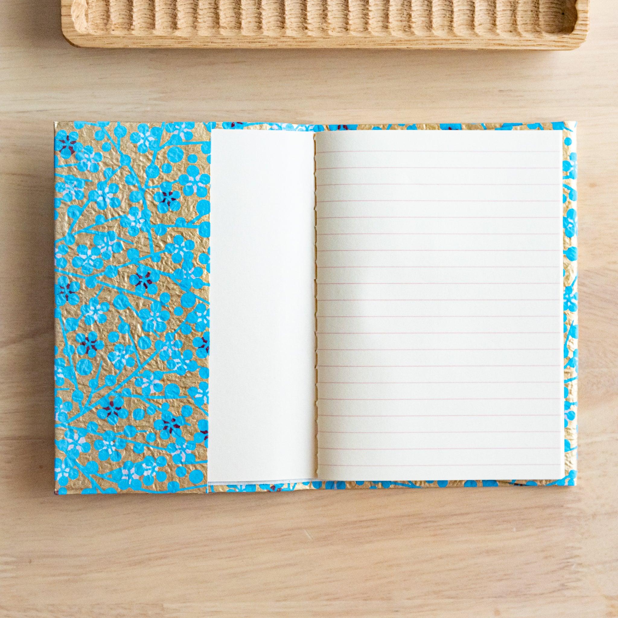 An open notebook with gold and blue floral patterned covers, showing one lined page and a blank page on a wooden surface.