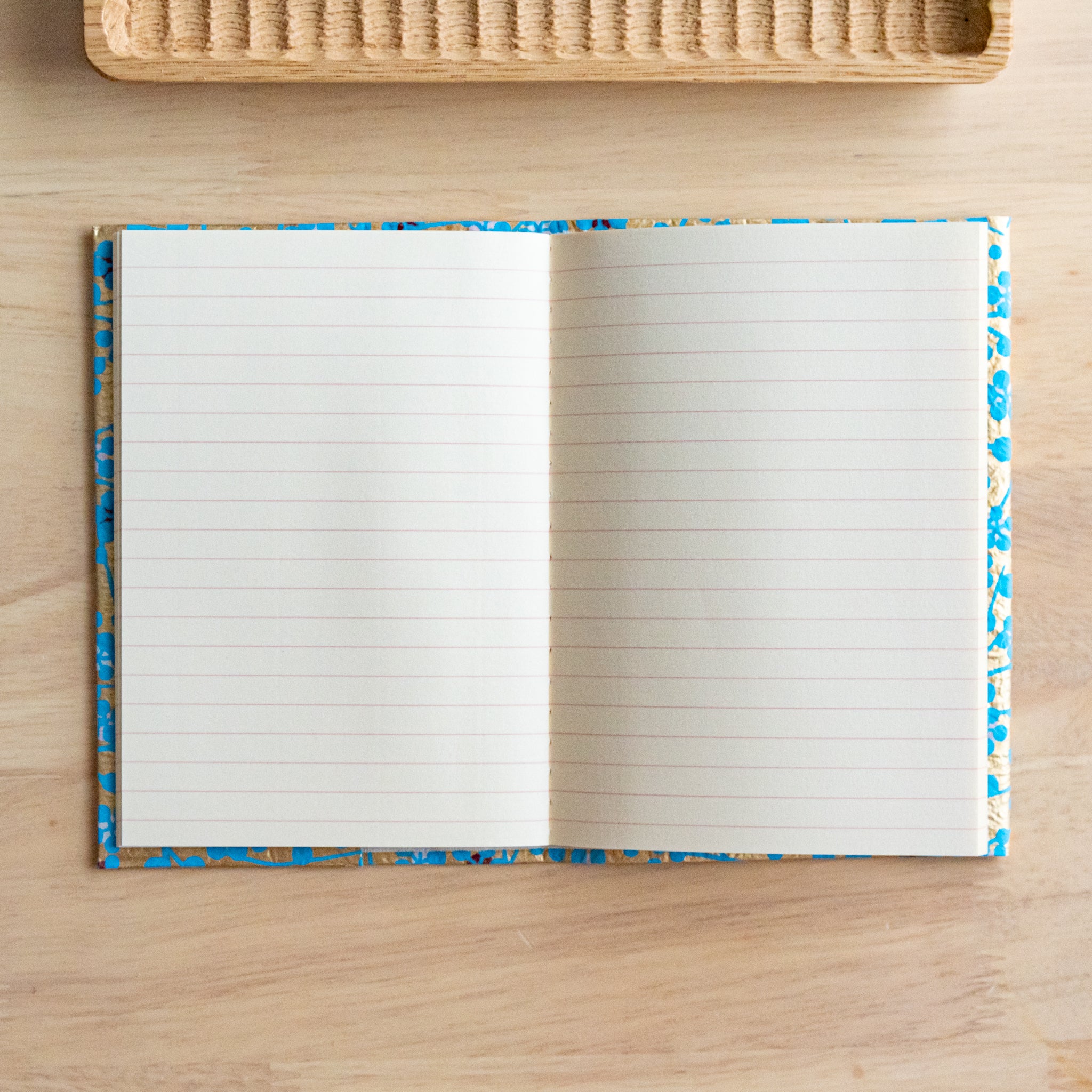 An open notebook with gold and blue floral covers, fully revealing two lined pages, placed on a light wooden surface.