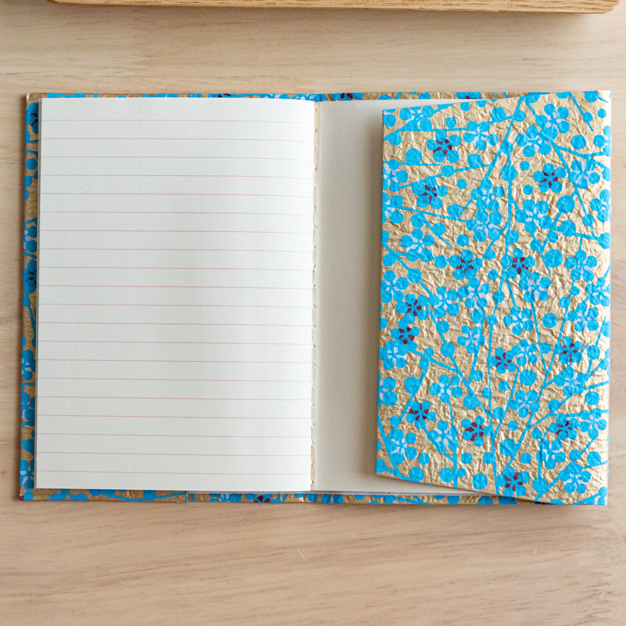 An open notebook with gold and blue floral covers, showing a lined page and an inner pocket on a wooden surface.