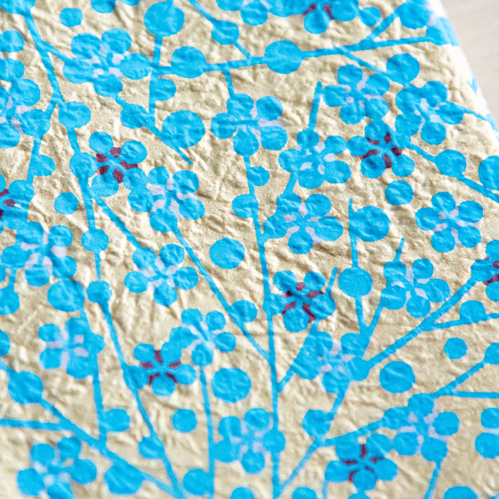 A close-up of a gold and blue floral pattern, showing intricate details of flowers and branches on textured paper.