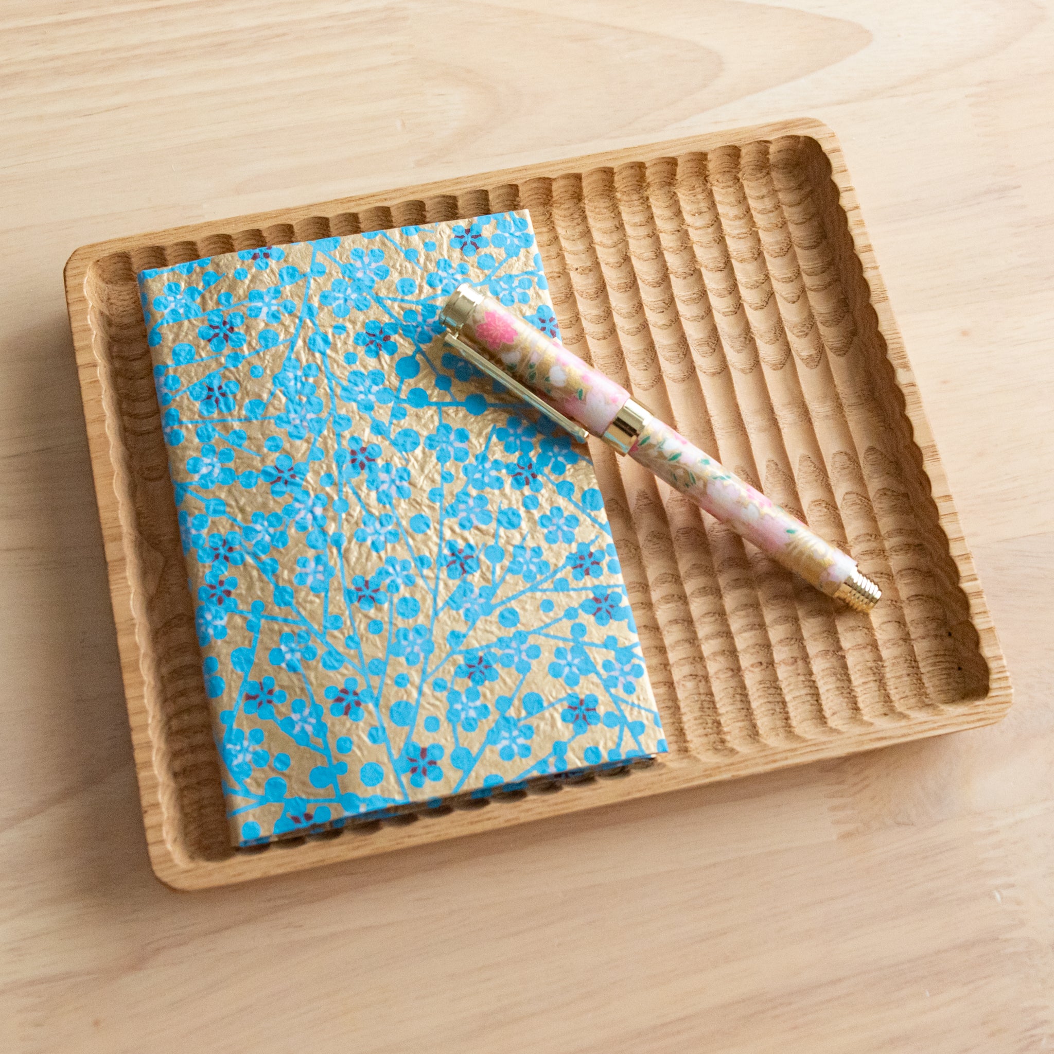 A gold and blue floral patterned notebook with a matching pink floral pen, placed on a wooden tray on a light wooden surface.