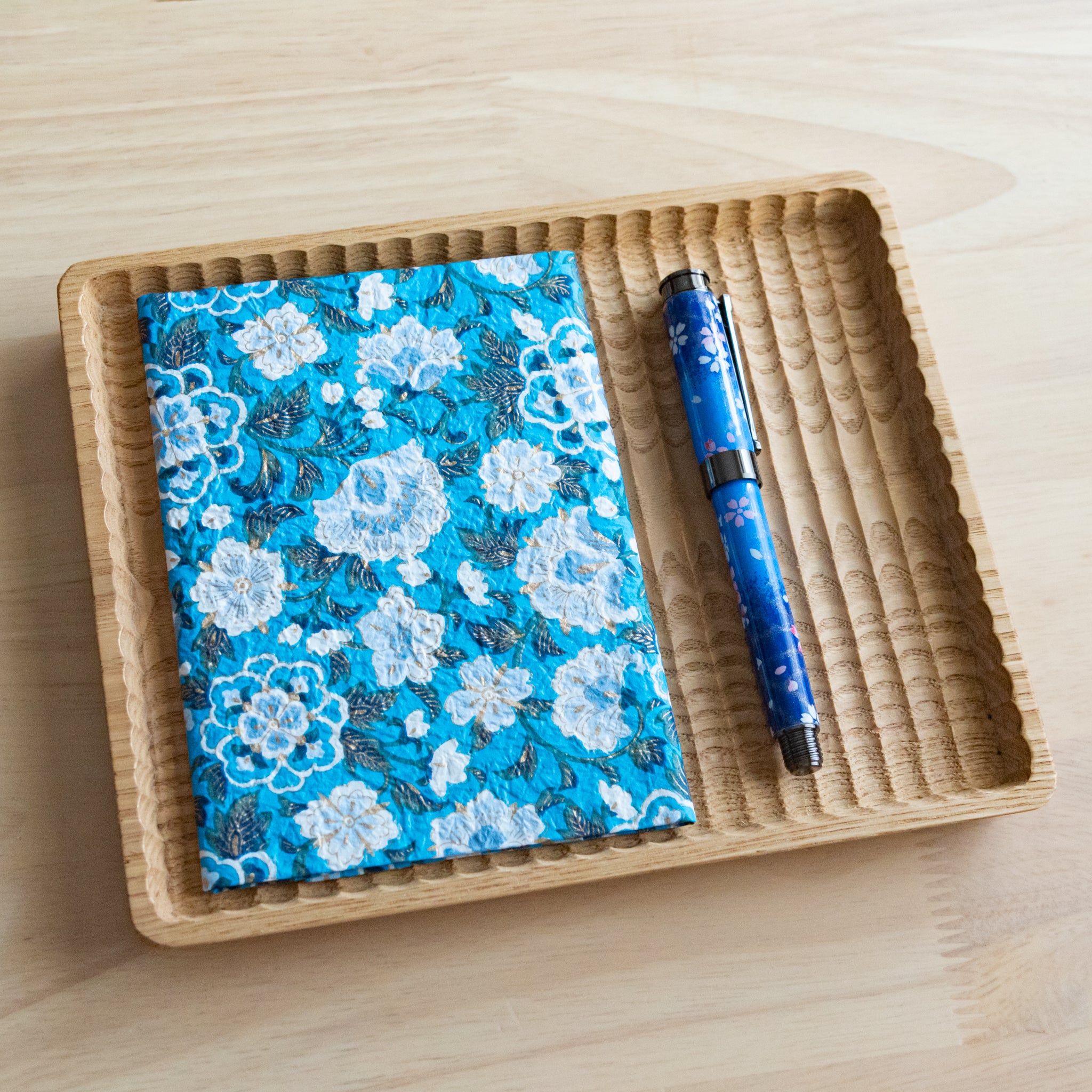 A blue floral patterned notebook and a matching blue pen, placed on a wooden tray on a light wooden surface.