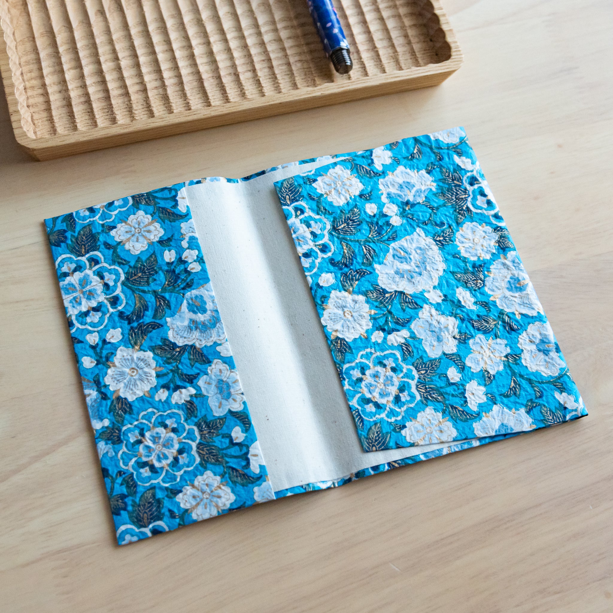 An open notebook cover with a blue floral pattern featuring white flowers and gold accents, showing its inner white lining.