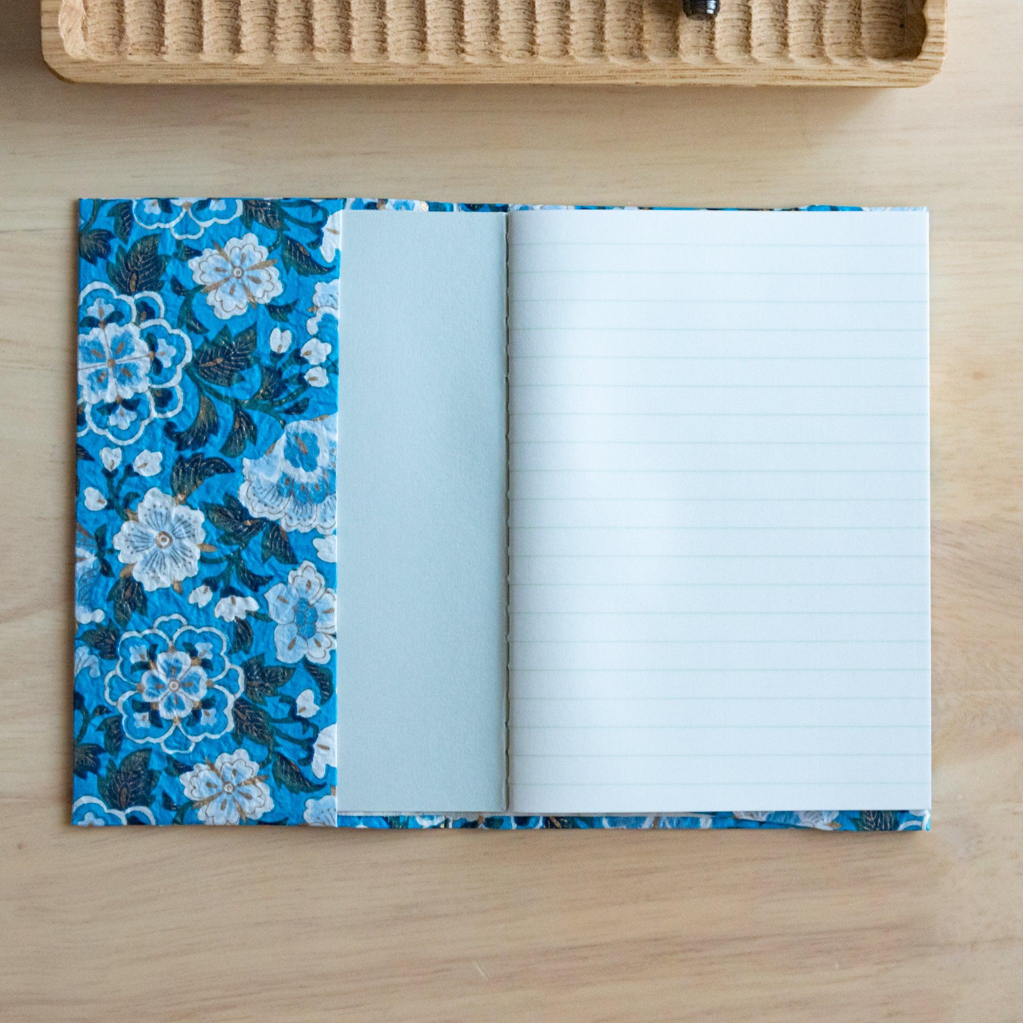 An open notebook with blue floral patterned covers, revealing one blank page and one lined page, placed on a wooden surface.