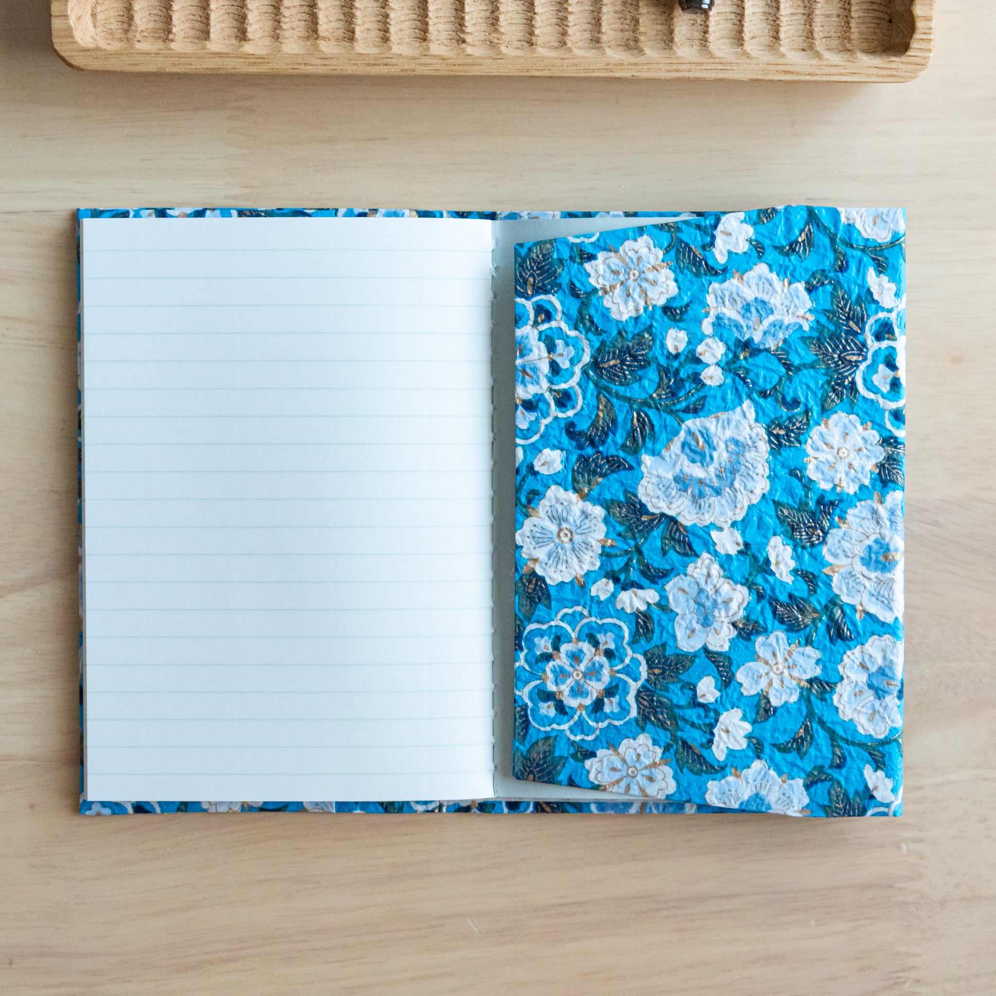 An open notebook with a blue floral pattern, showing a lined page and an inner pocket, placed on a light wooden surface.