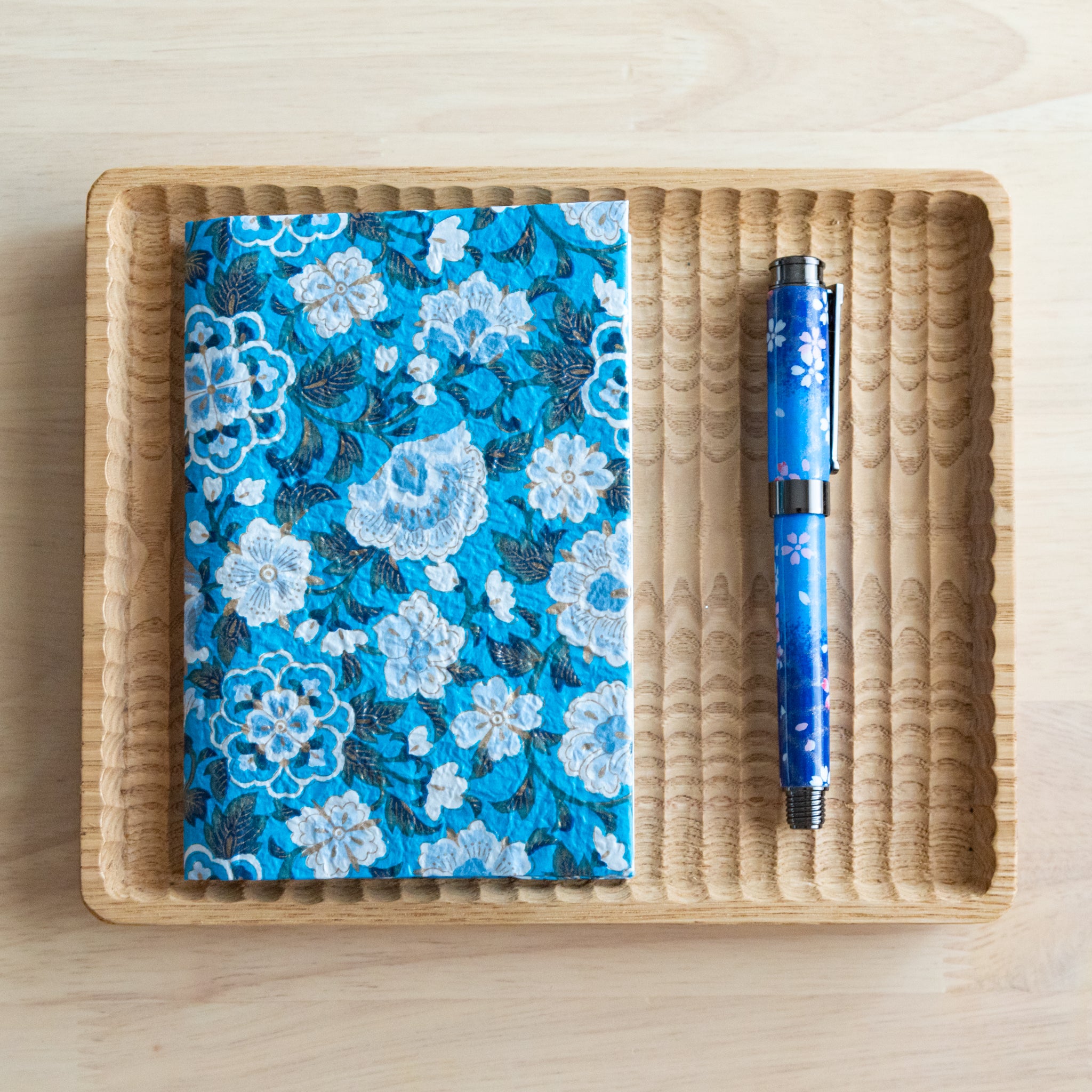 A blue floral patterned notebook and a matching blue pen arranged neatly on a wooden tray on a light wooden surface.