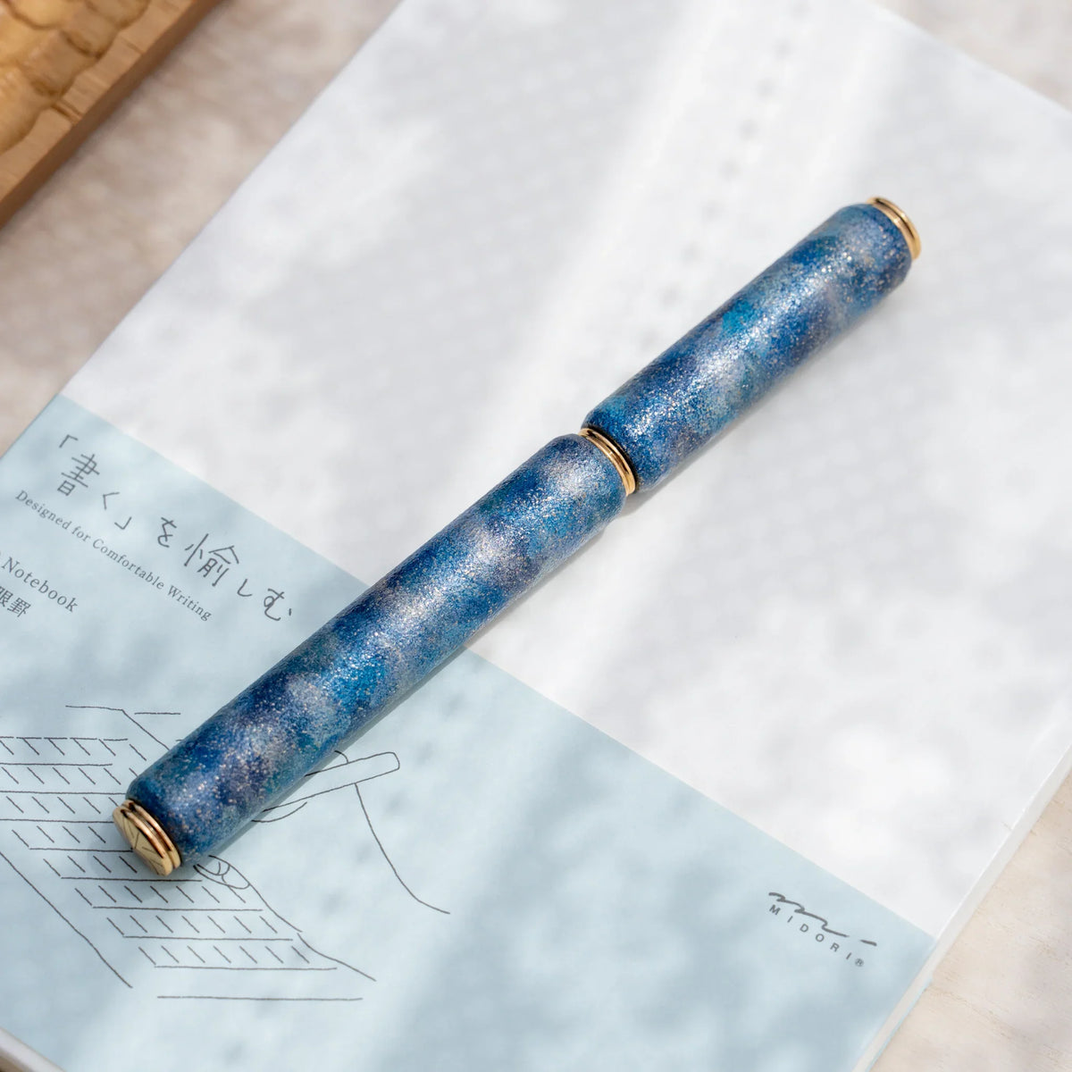 Blue marbled urushi lacquered rollerball pen in zelkova wood, resting on a Japanese notebook