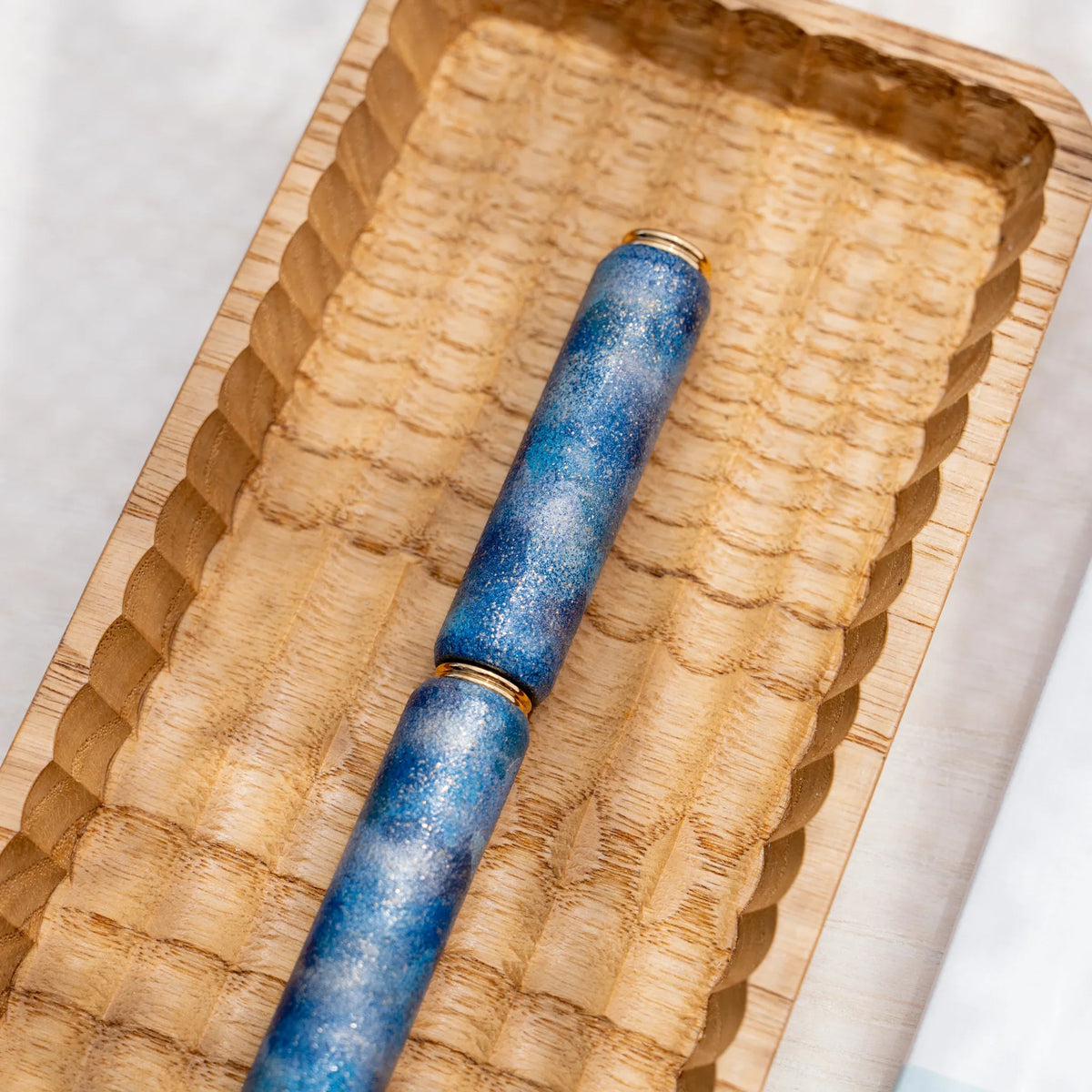 Blue marbled urushi lacquered rollerball pen in zelkova wood, angled view in a carved wooden tray
