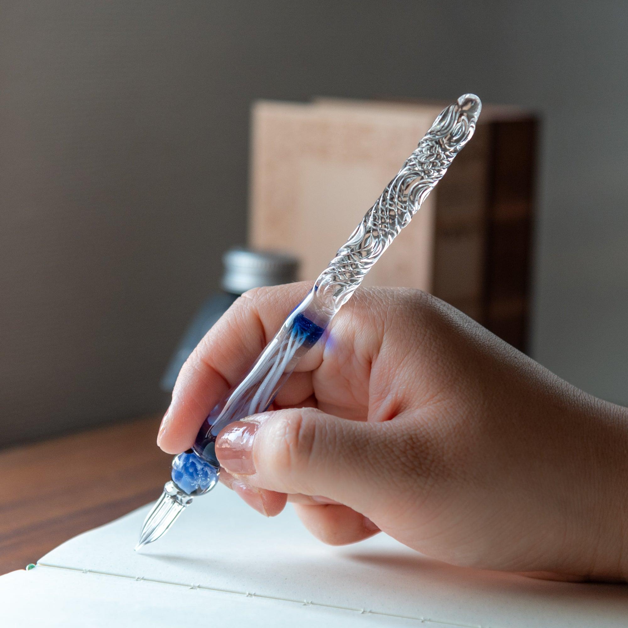 A hand holding a glass pen with an blue rose motif, writing on a notebook, capturing the elegance of the pen in use.