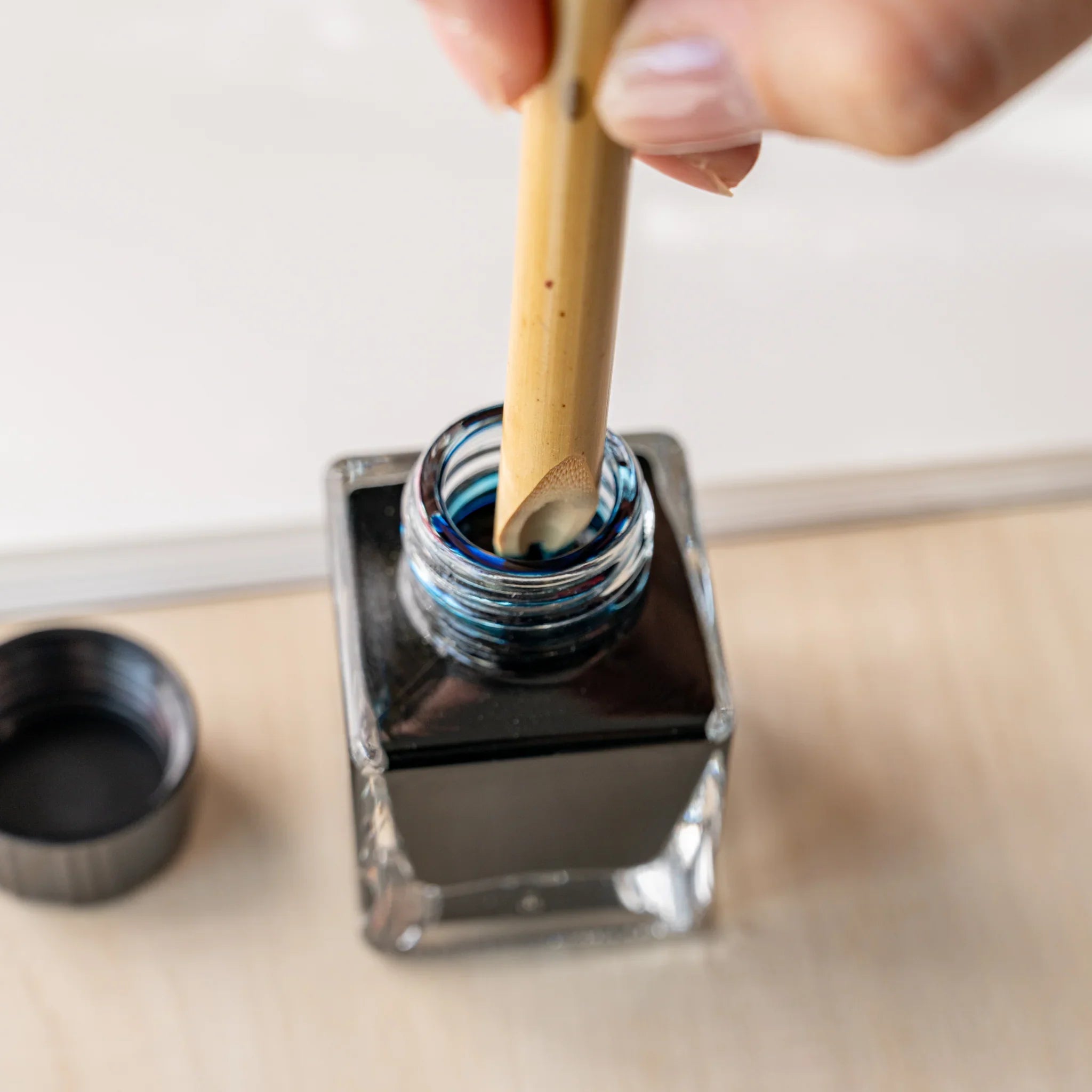 Hand lowering Boku-Undo bamboo pen nib into ink bottle filled with dark blue-black ink, open bottle cap at left