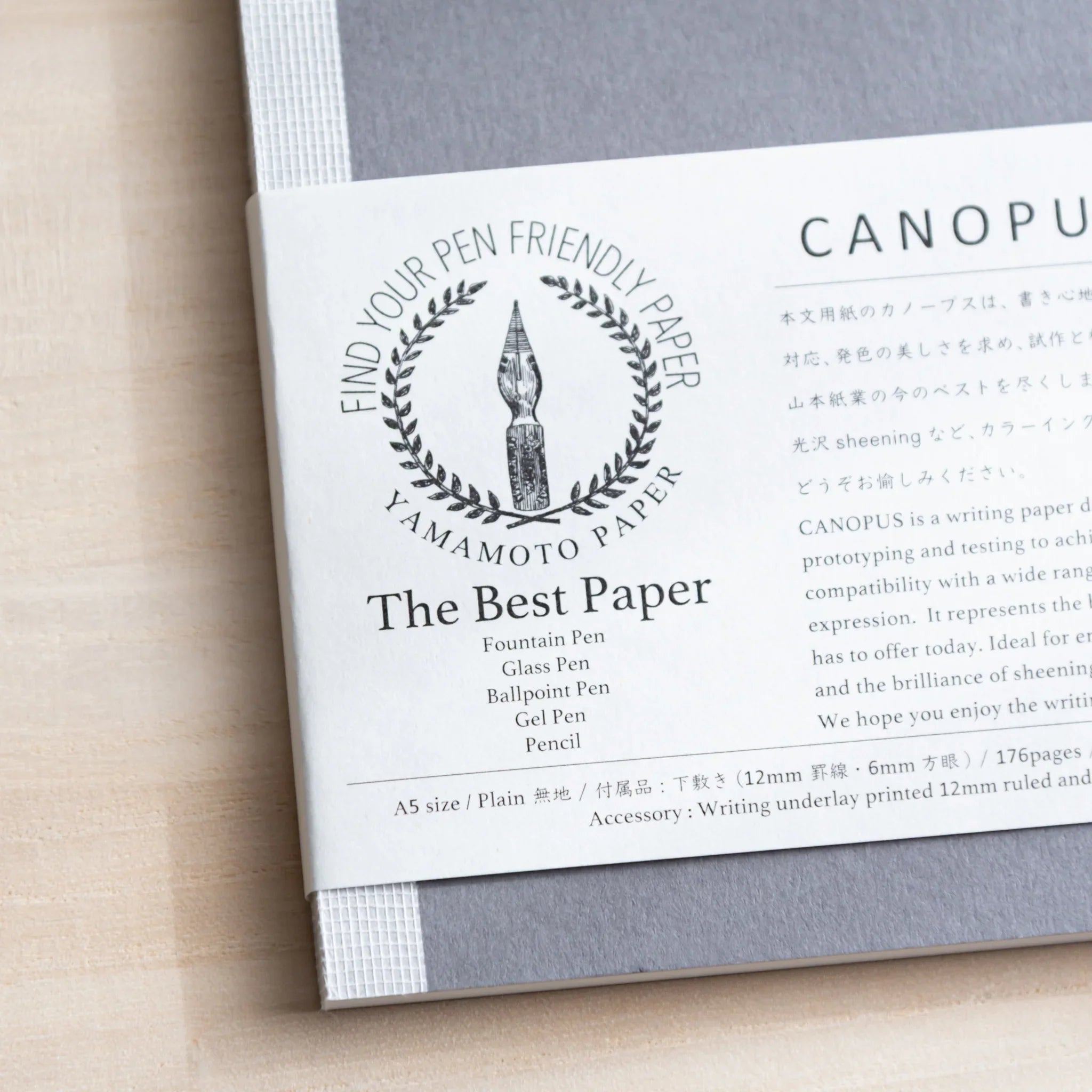 Close-up of grey Japanese Notebook showing white label with 'CANOPUS' and 'The Best Paper' on light wooden desk.
