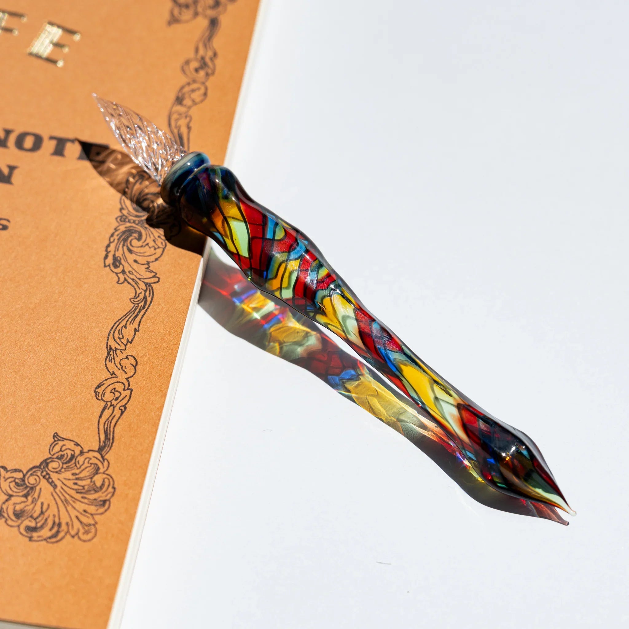 Glass dip pen with a clear twisted nib and stained-glass mosaic body in red, yellow, blue and green, on a kraft notebook
