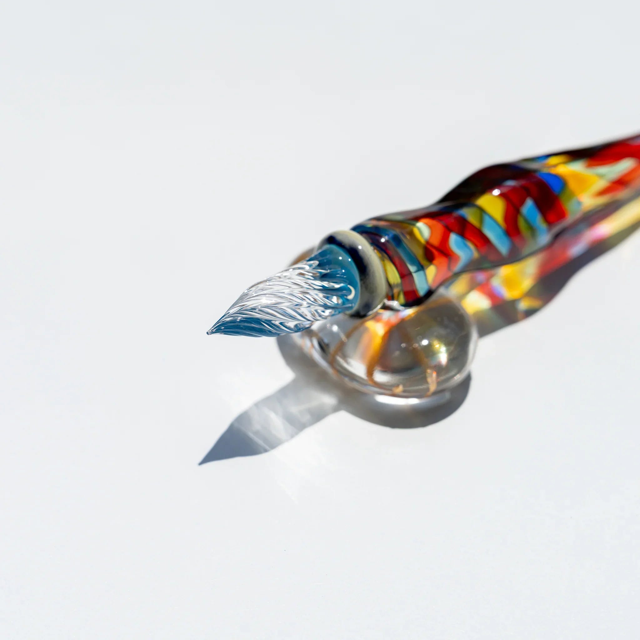 Angled view of the Cathedral glass dip pen nib, clear grooved tip with blue collar, mosaic barrel resting on a glass rest