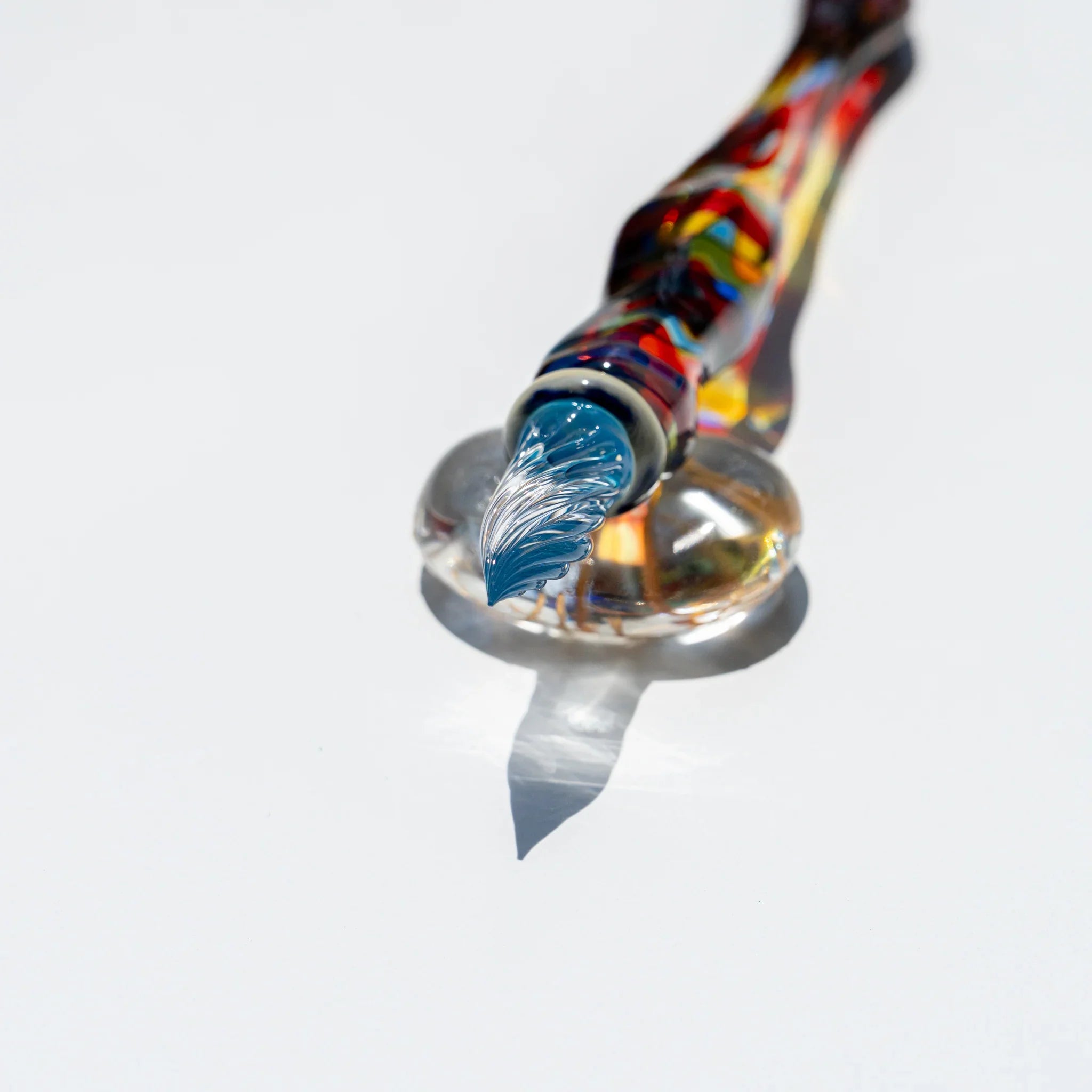 Cathedral glass dip pen resting nib-down on a clear glass rest, blue tinted grooved nib, red, yellow and blue mosaic barrel