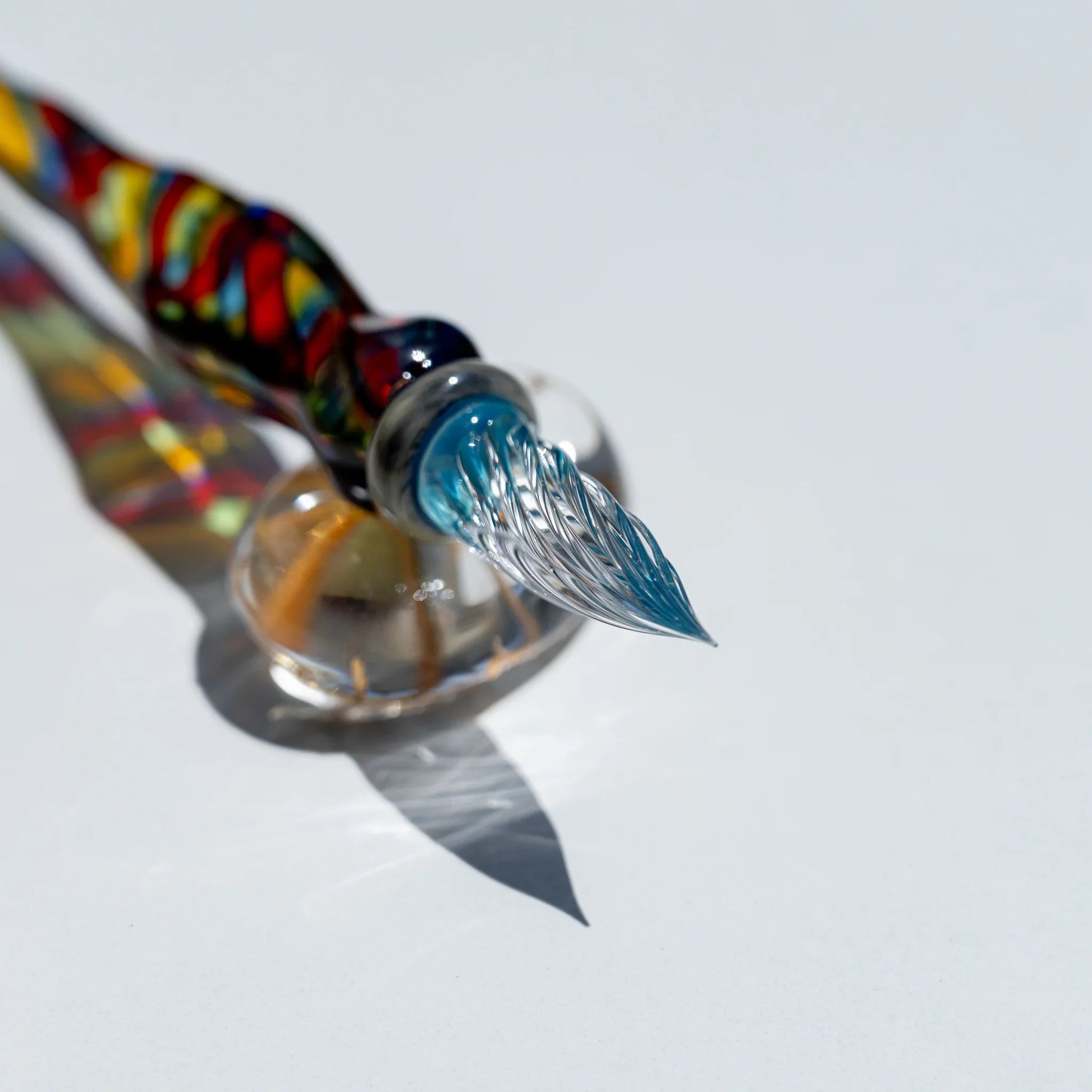 Glass dip pen nib end with aqua tip, clear ribbed nib resting on a clear round glass rest on white