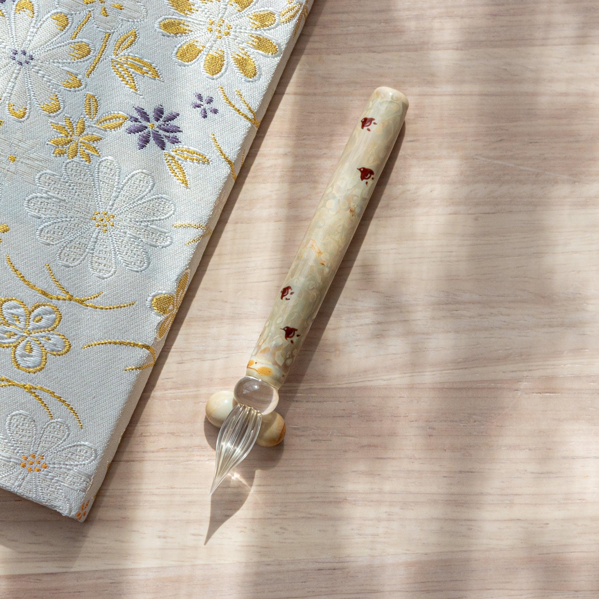 Ivory glass pen with Chidori bird design, paired with a matching glass pen rest. Displayed on a wooden surface beside a Nishijin textile notebook.