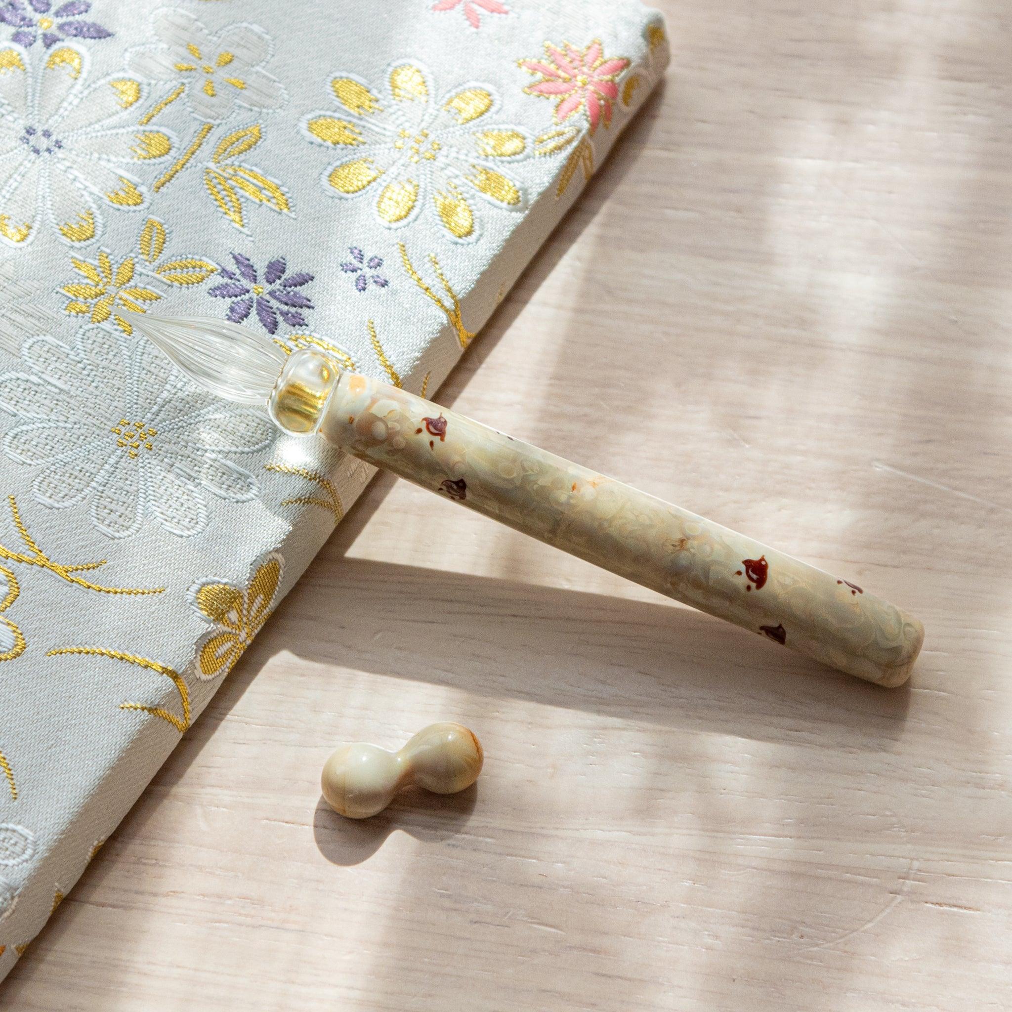 Ivory glass pen with Chidori bird design, paired with a matching glass pen rest. Displayed on a wooden surface beside a Nishijin textile notebook.