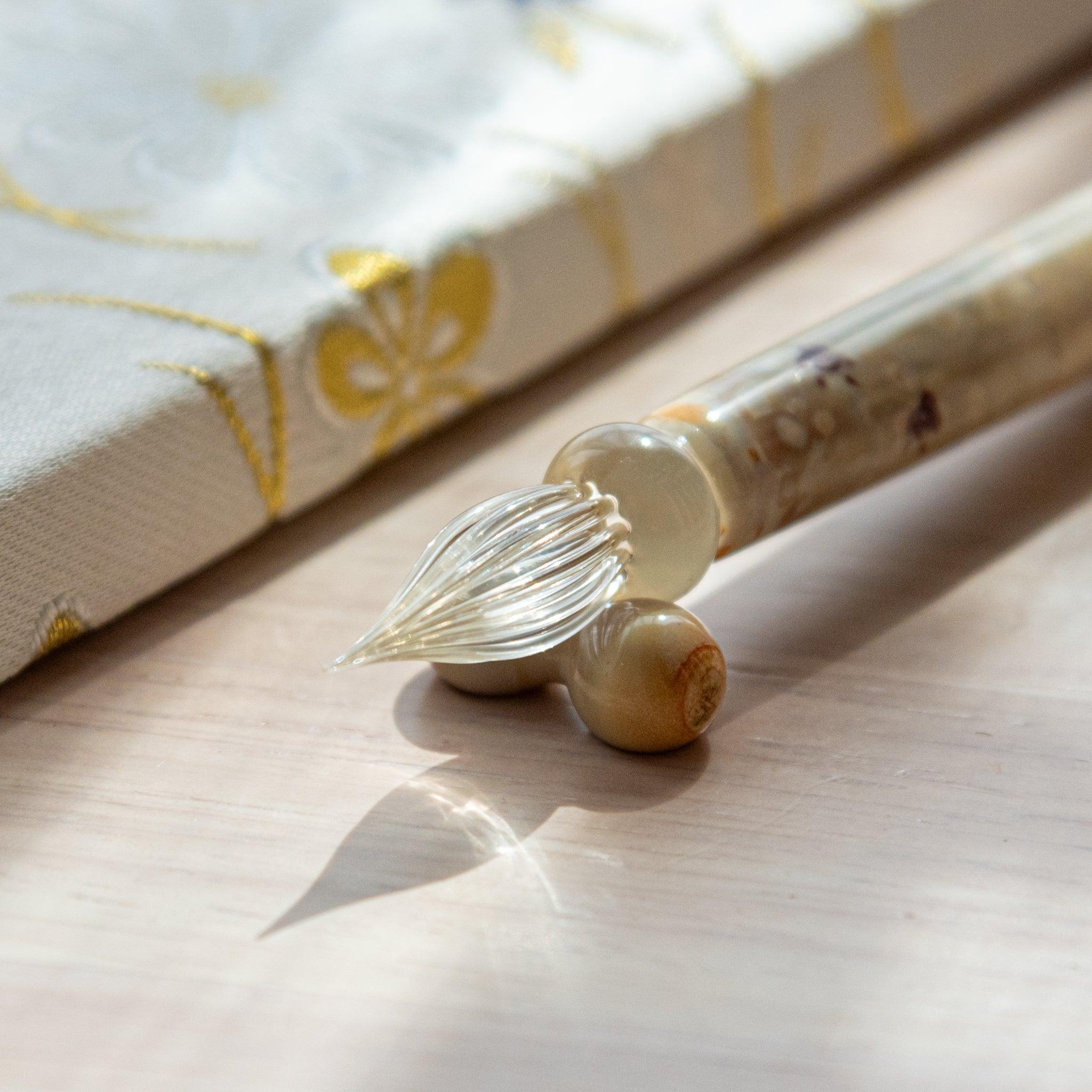 Ivory glass pen with Chidori bird design, paired with a matching glass pen rest. Displayed on a wooden surface beside a Nishijin textile notebook.