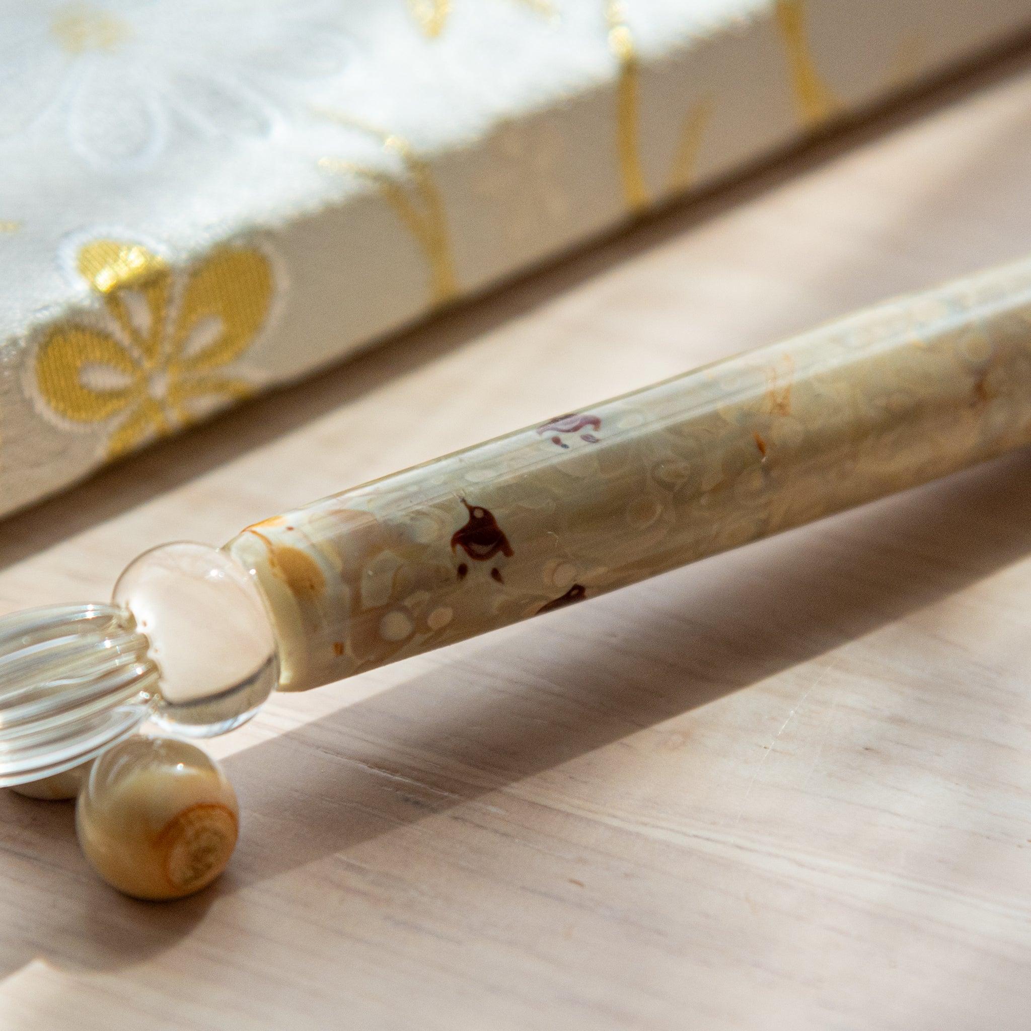 Ivory glass pen with Chidori bird design, paired with a matching glass pen rest. Displayed on a wooden surface beside a Nishijin textile notebook.