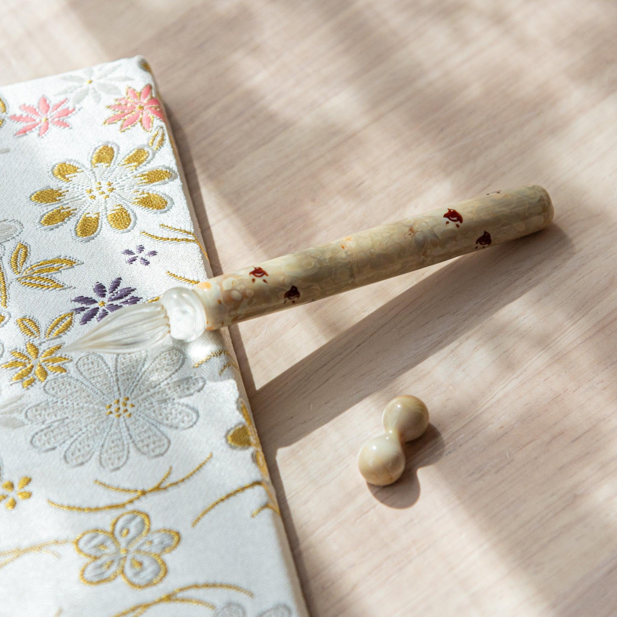 Ivory glass pen with Chidori bird design, paired with a matching glass pen rest. Displayed on a wooden surface beside a Nishijin textile notebook.