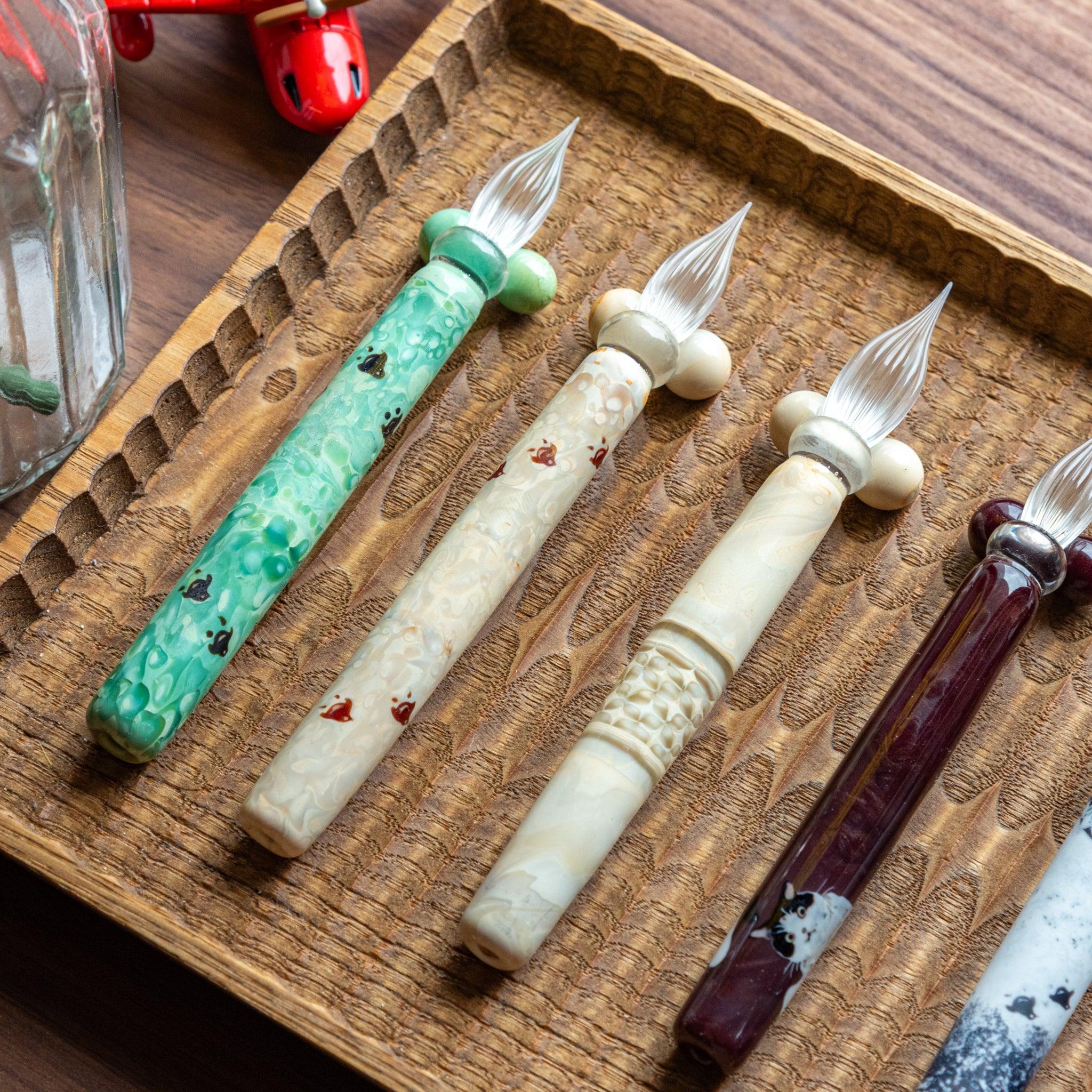 Four glass pens featuring traditional designs, including Chidori, arranged on a hand-carved wooden pen tray. Elegant and unique designs.