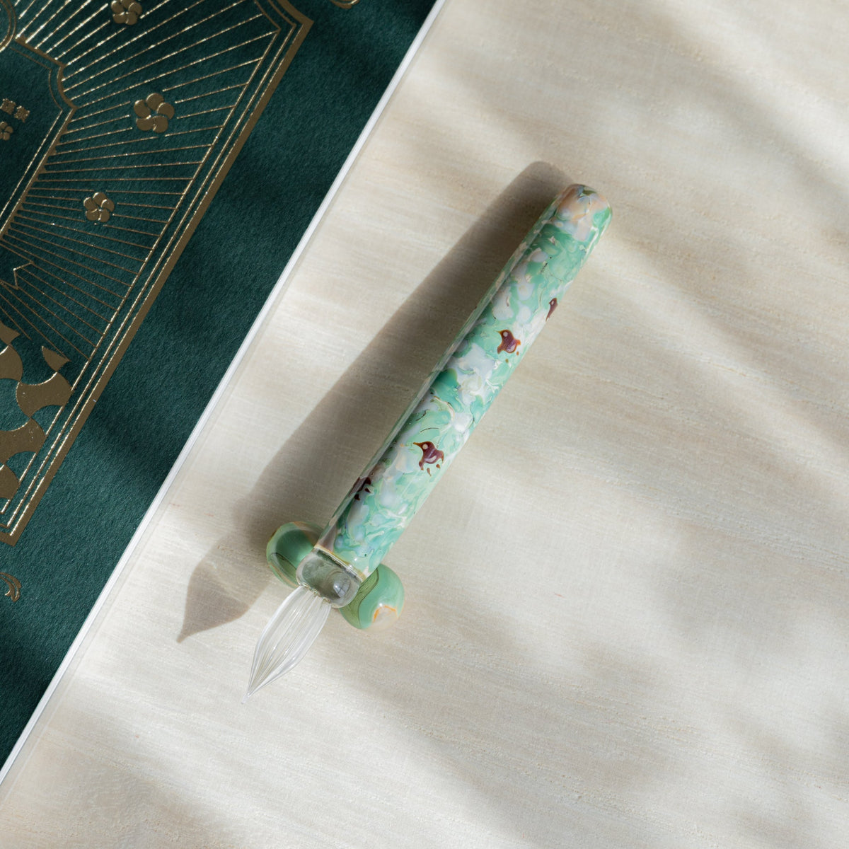 Chidori Mint Marble glass dip pen angled nib down beside a green notebook with gold foil printing