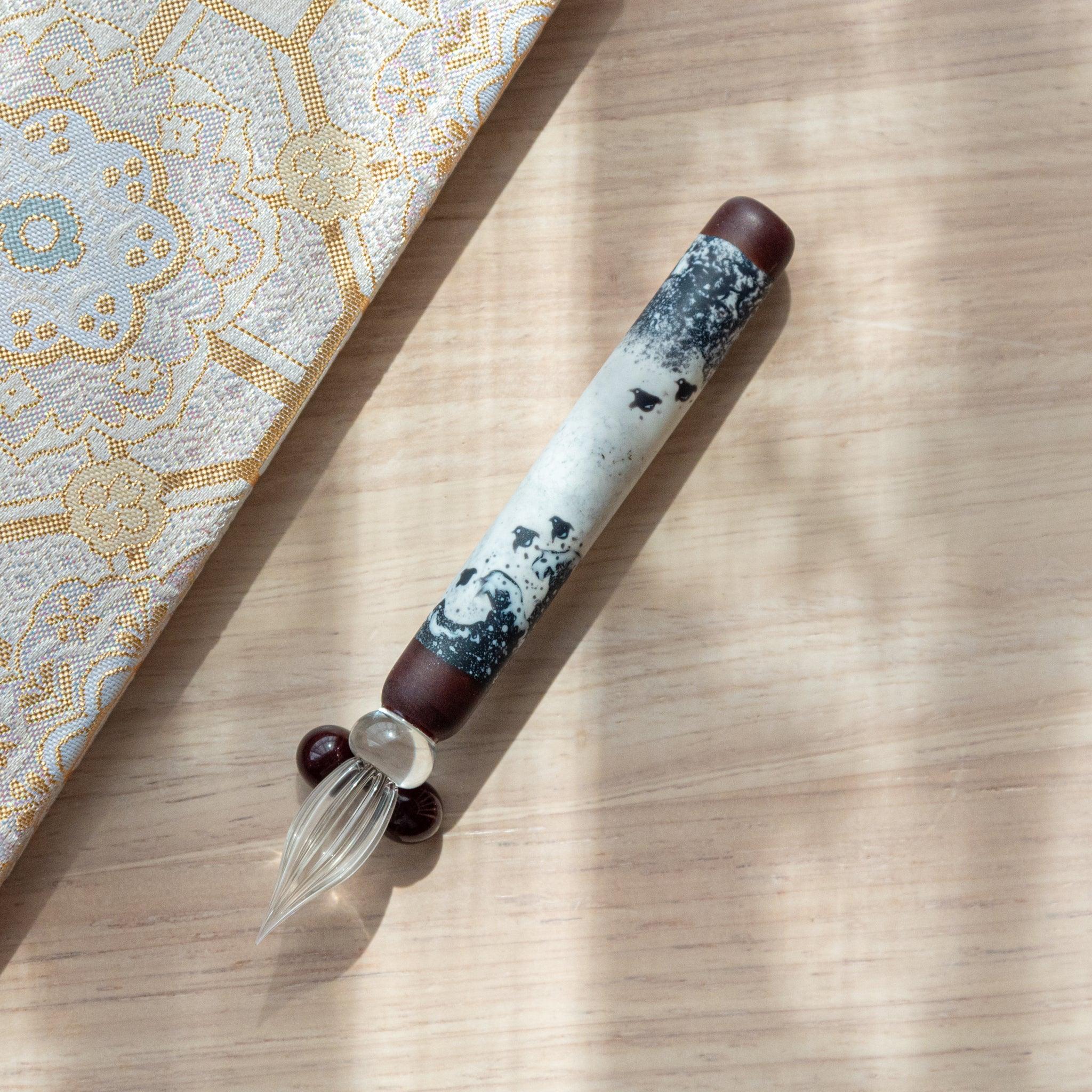 Black and white glass pen with a Chidori on Great Wave design, paired with a matching glass pen rest. Displayed on a wooden surface with a Nishijin textile notebook.