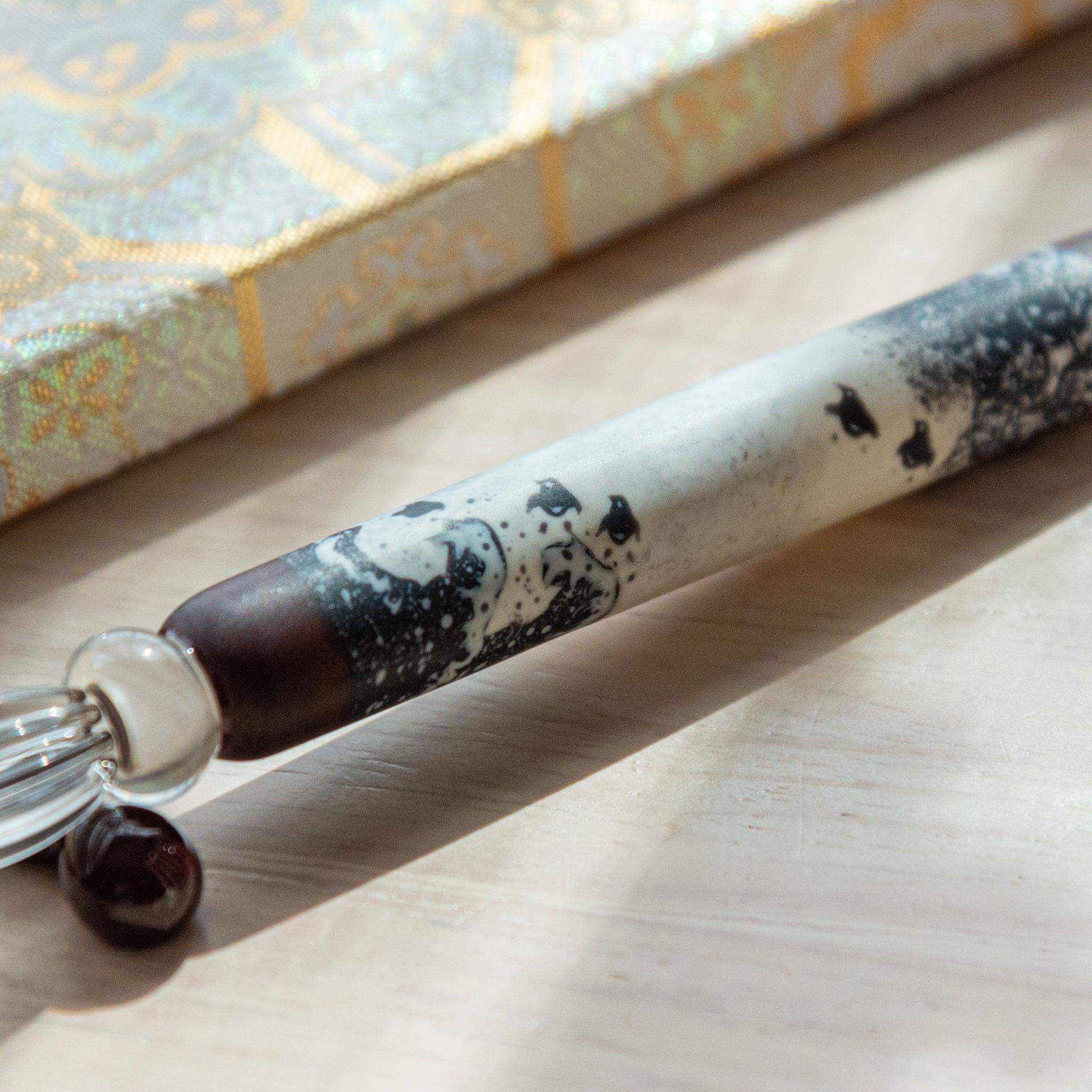 Black and white glass pen with a Chidori on Great Wave design, paired with a matching glass pen rest. Displayed on a wooden surface with a Nishijin textile notebook.