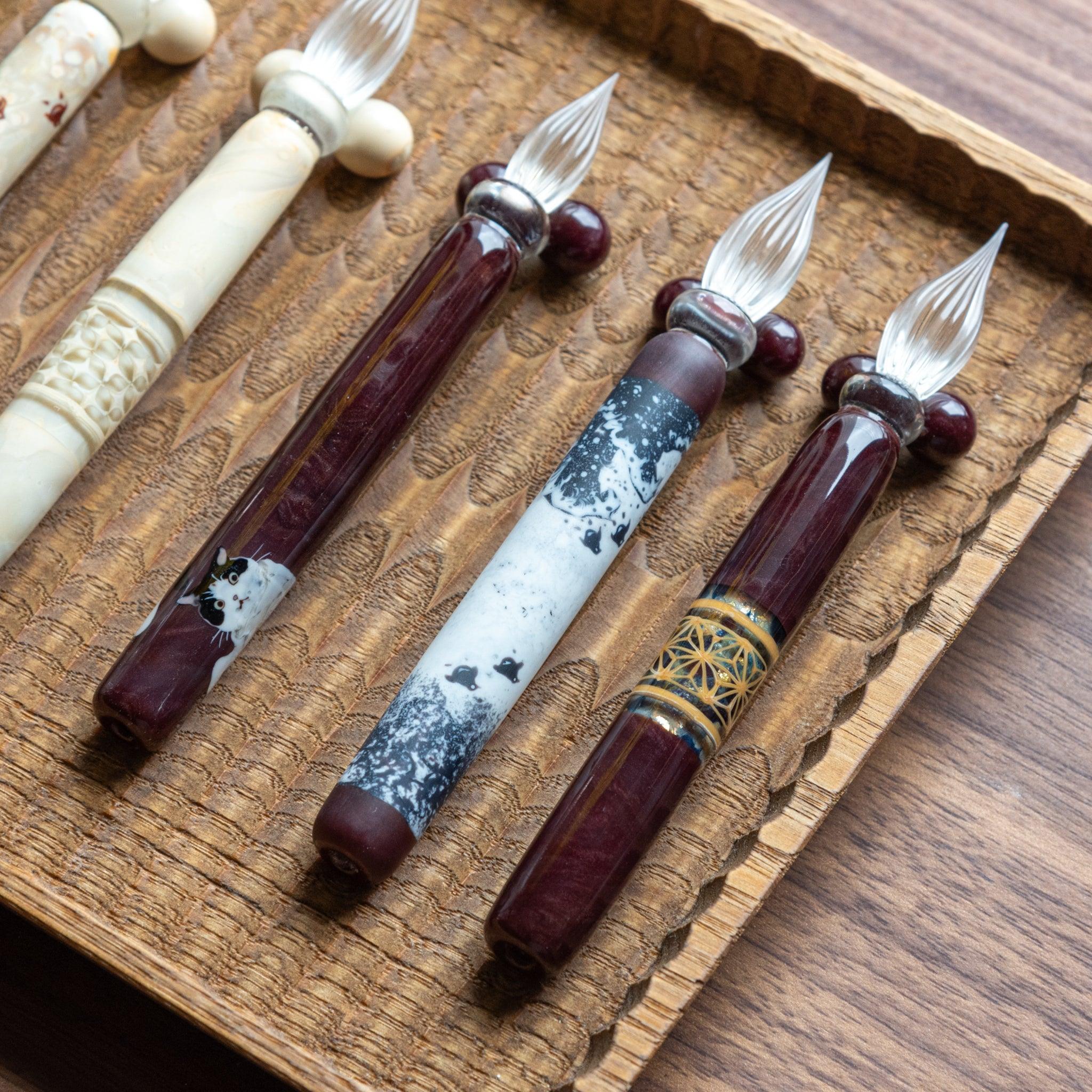 Four glass pens featuring traditional designs, including Chidori, arranged on a hand-carved wooden pen tray. Elegant and unique designs.