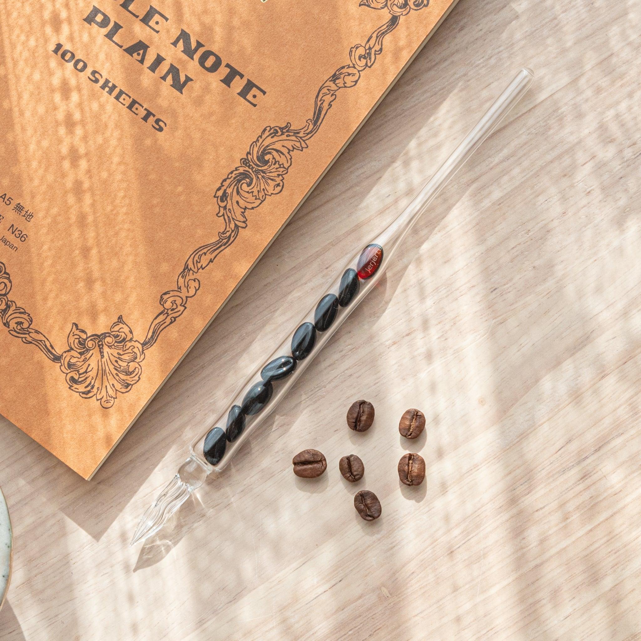 A glass dip pen with coffee bean-shaped glass pieces inside its shaft rests on a wooden surface near coffee beans and a plain notebook.