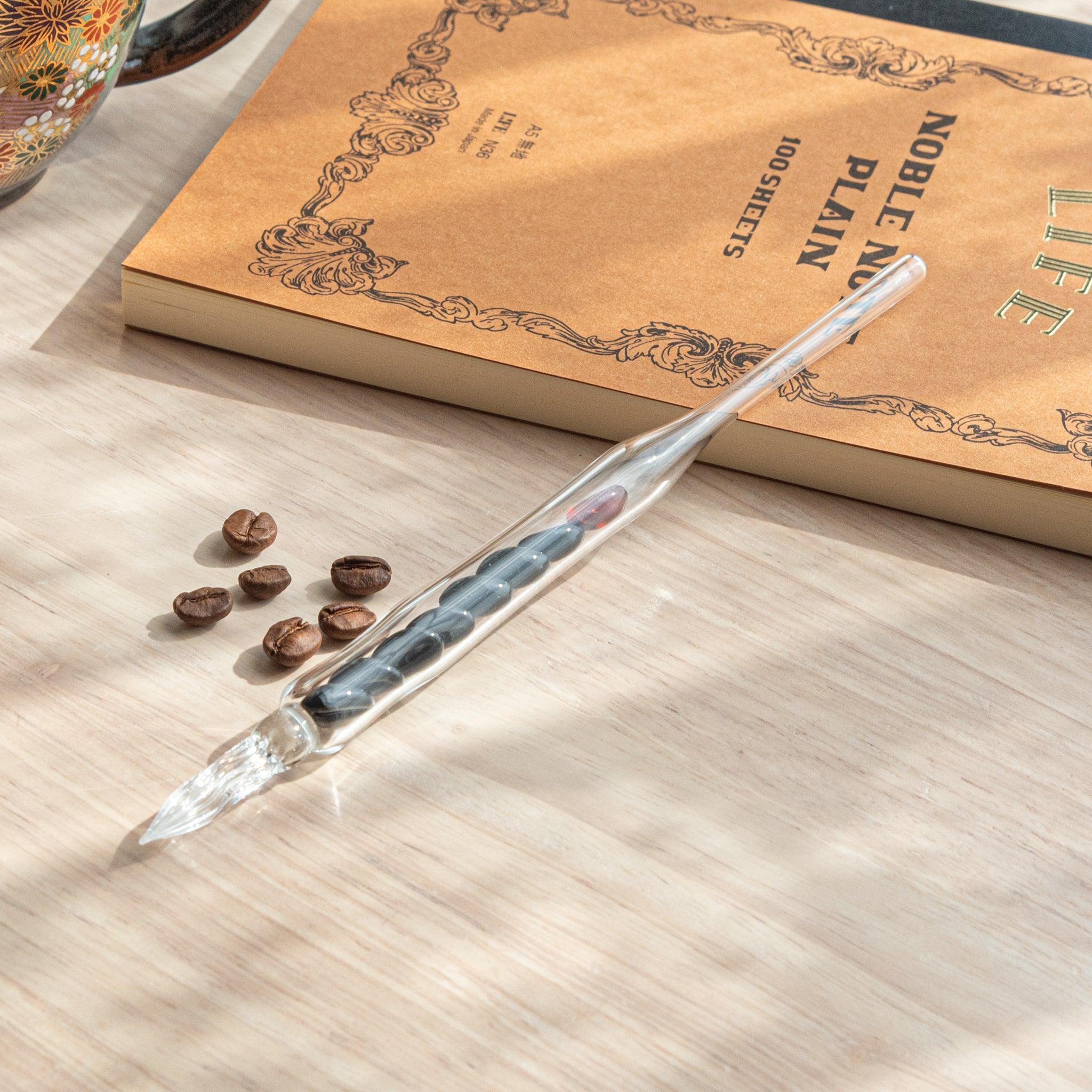 A glass dip pen with coffee bean-shaped glass pieces inside its shaft rests on a wooden surface near coffee beans and a plain notebook.