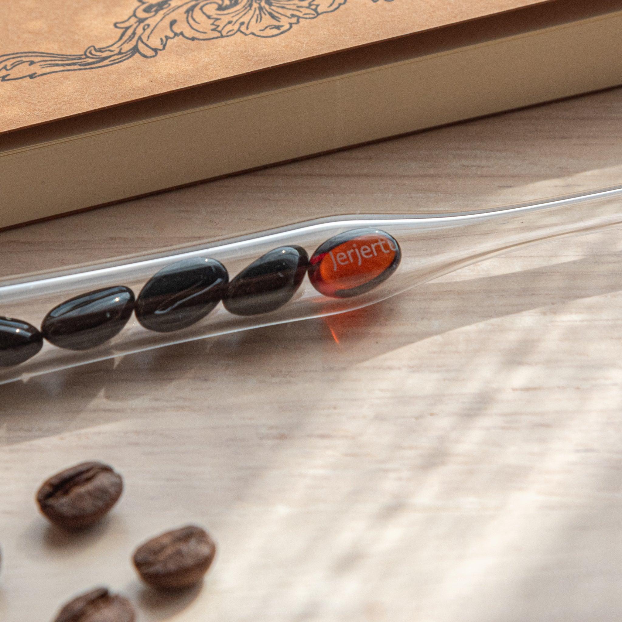 A glass dip pen with coffee bean-shaped glass pieces inside its shaft rests on a wooden surface near coffee beans and a plain notebook.