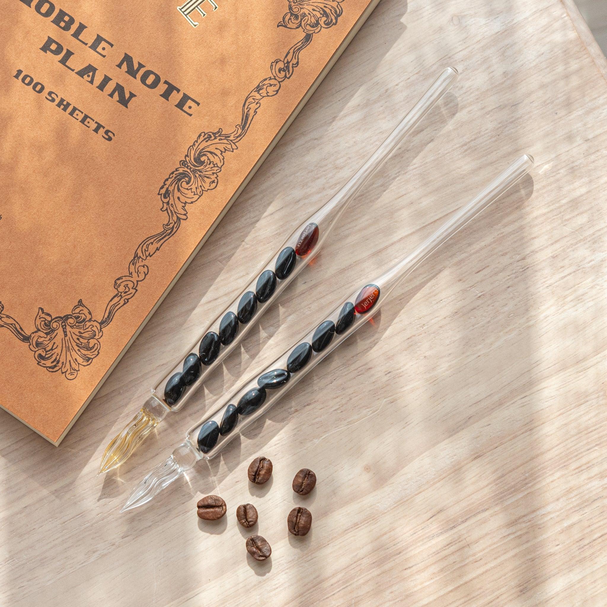 Two glass dip pens with coffee bean-shaped glass pieces inside their shafts are placed on a wooden surface near coffee beans and a plain notebook.