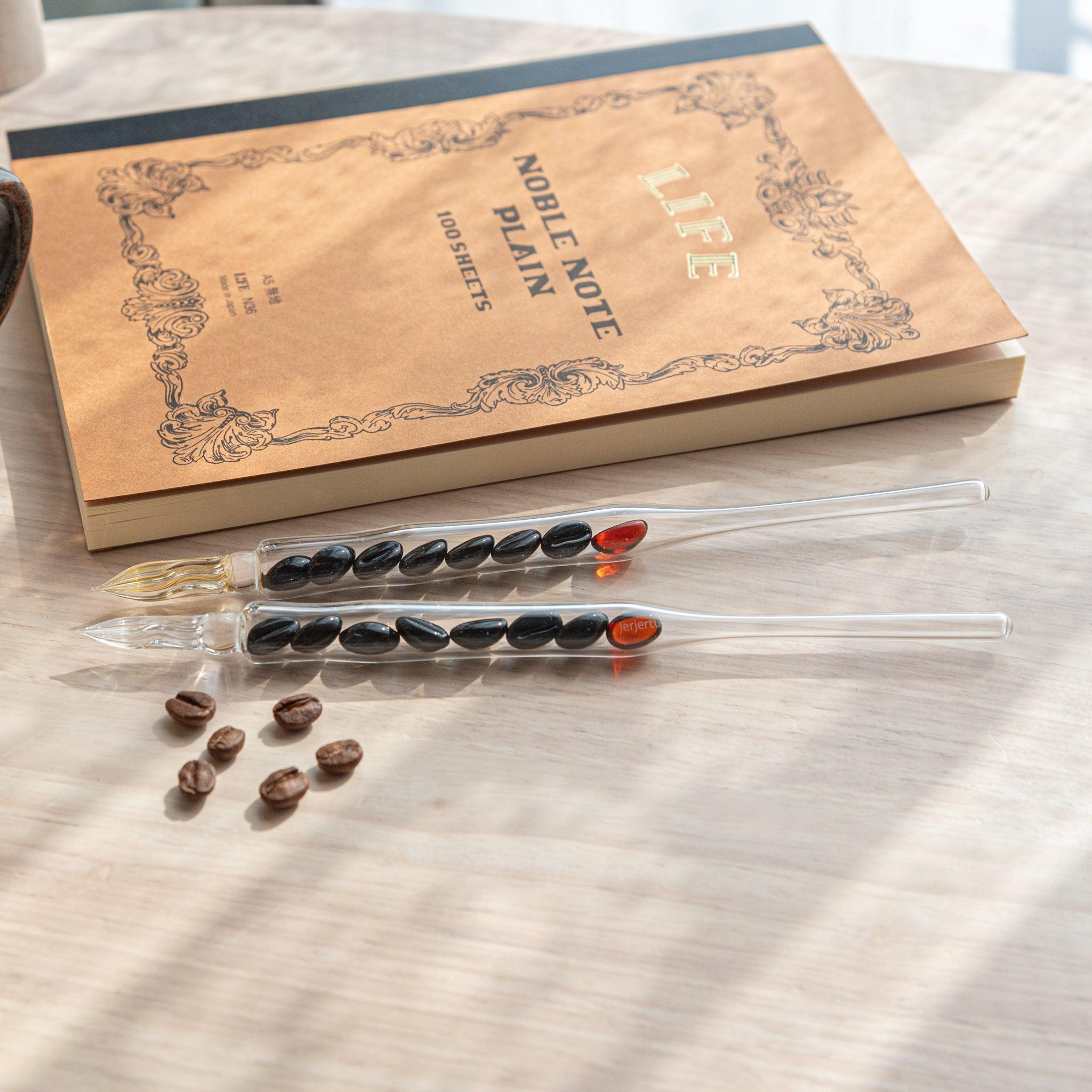 Two glass dip pens with coffee bean-shaped glass pieces inside their shafts are placed on a wooden surface near coffee beans and a plain notebook.