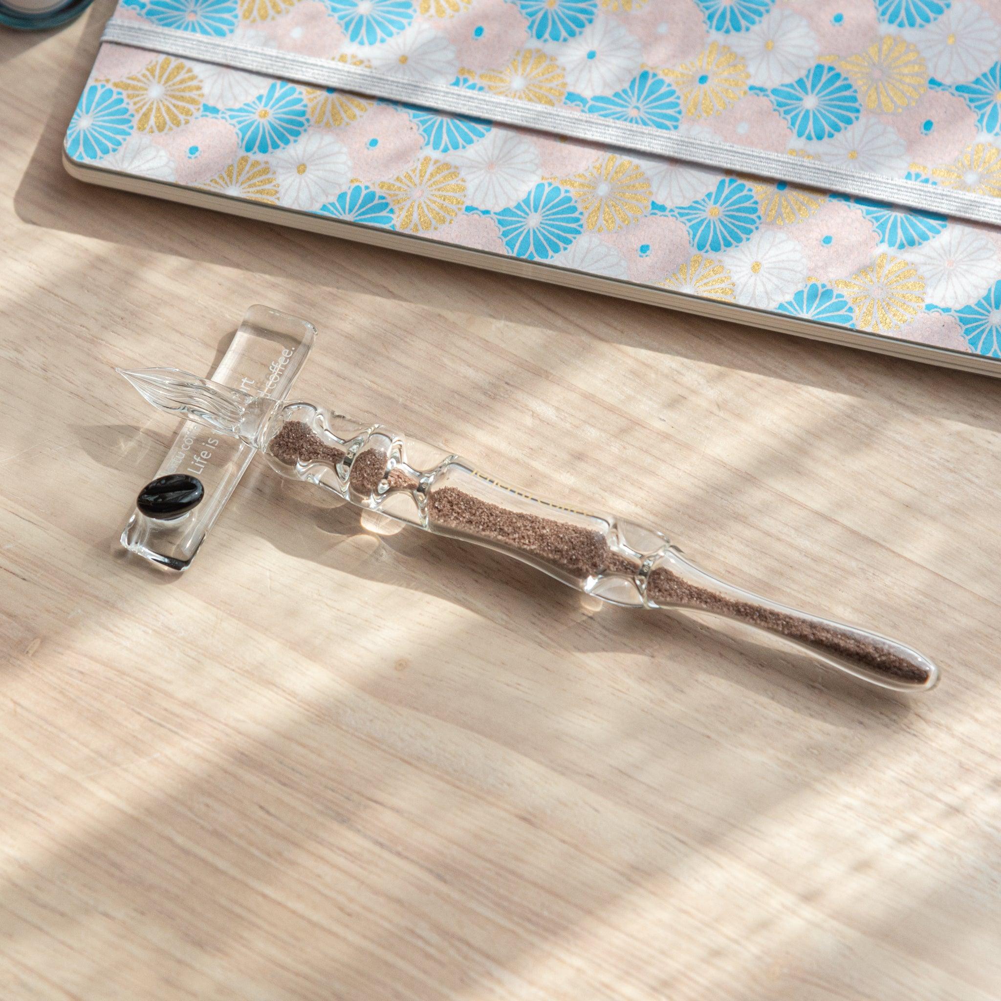 A glass dip pen with brown sand-like particles inside its shaft is resting on a glass pen rest. The pen is placed on a wooden surface near a notebook with pastel floral patterns.