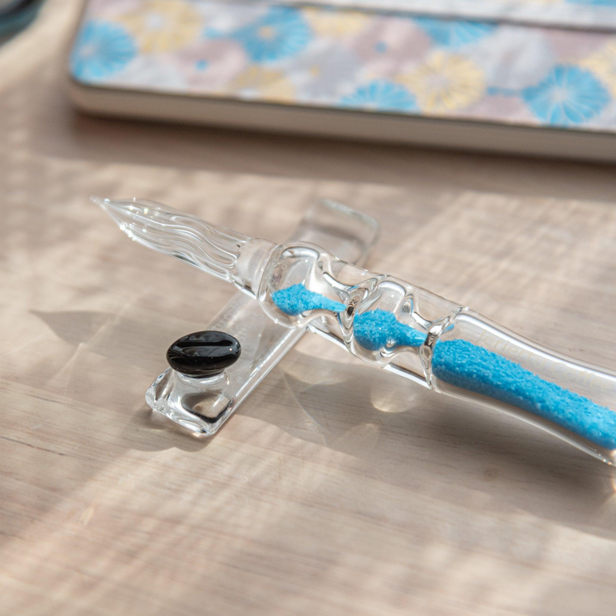 Close-up of a glass dip pen with blue sand-like particles inside its shaft, resting on a glass rest. The background features a notebook with pastel floral patterns.