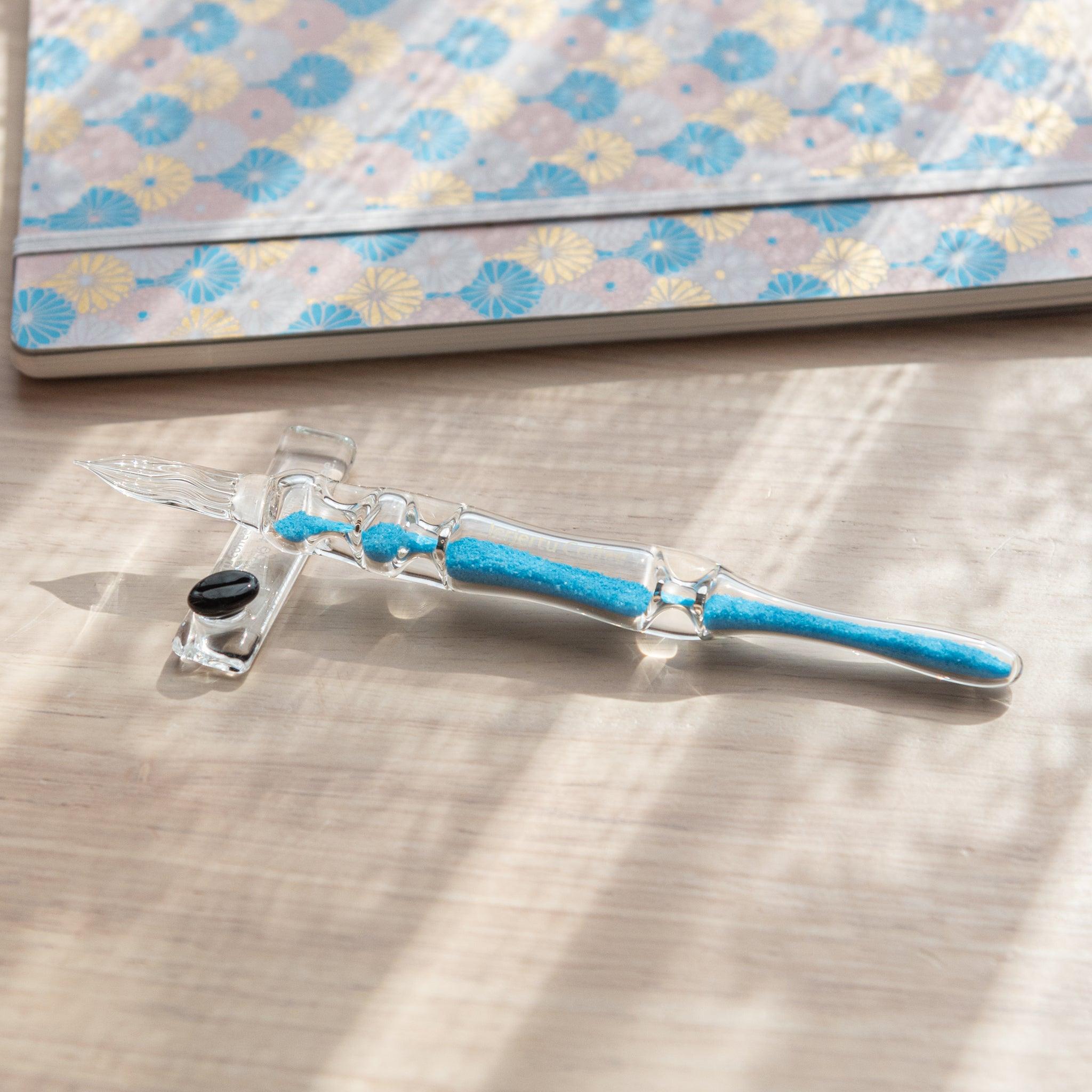 A glass dip pen with blue sand-like particles inside its shaft is resting on a glass pen rest. The pen is placed on a wooden surface near a patterned notebook with pastel floral designs.