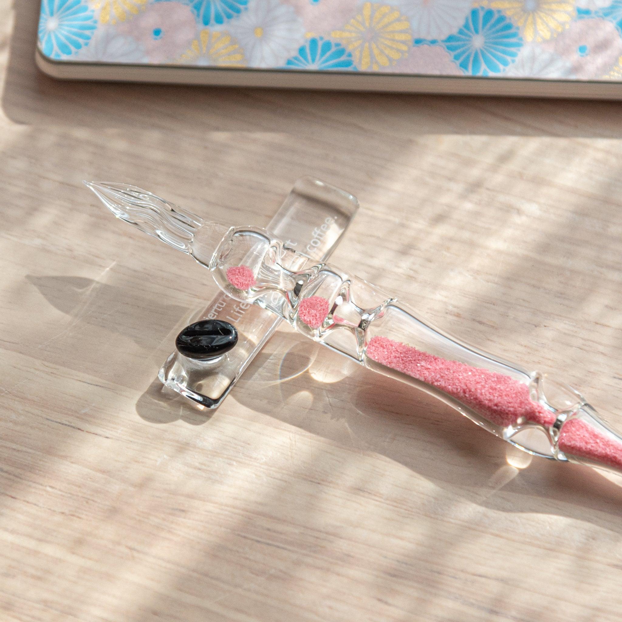 Close-up of a glass dip pen with pink sand-like particles inside its shaft, resting on a glass rest. The background shows part of a patterned notebook with floral designs.