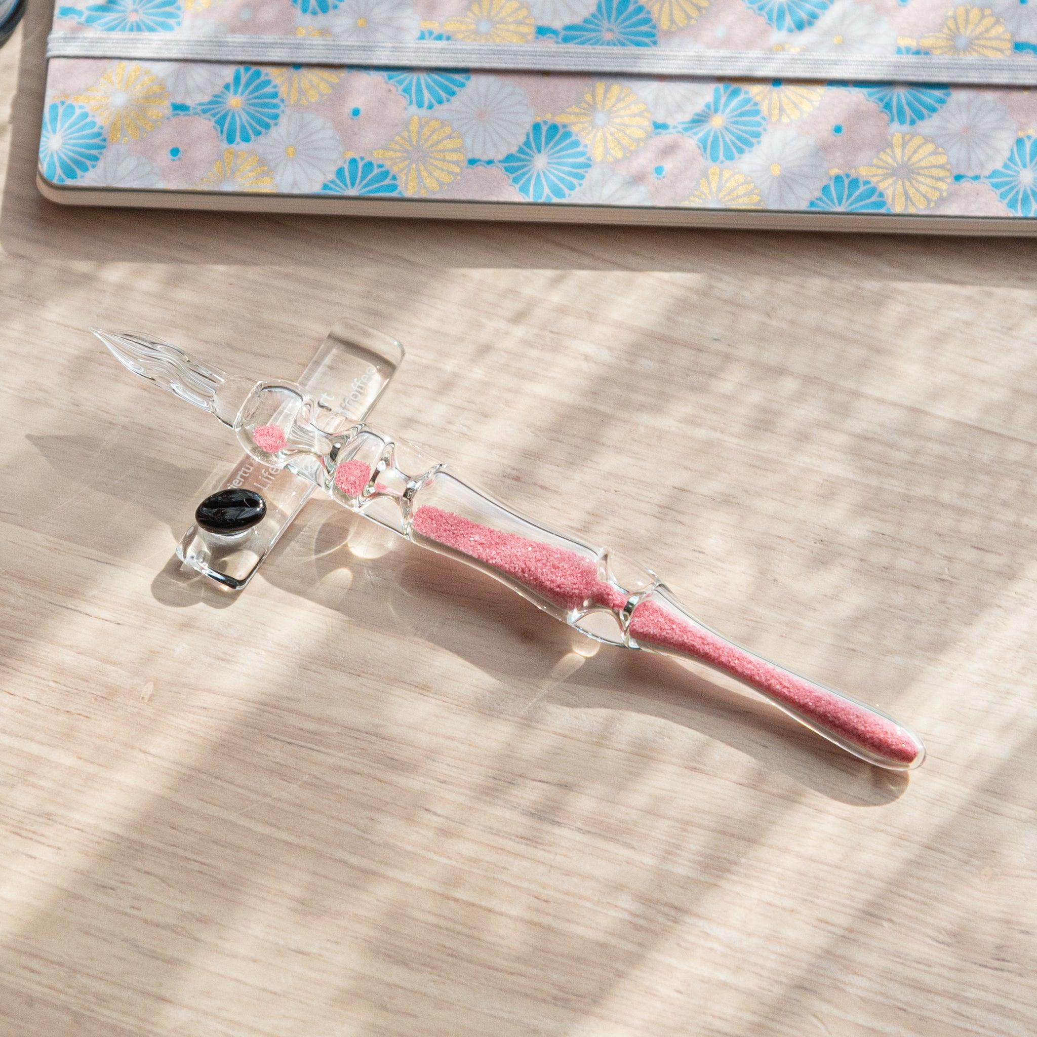 A glass dip pen with pink sand-like particles inside its shaft is resting on a glass pen rest. The pen is placed on a wooden surface near a patterned notebook with floral designs in pastel colors.